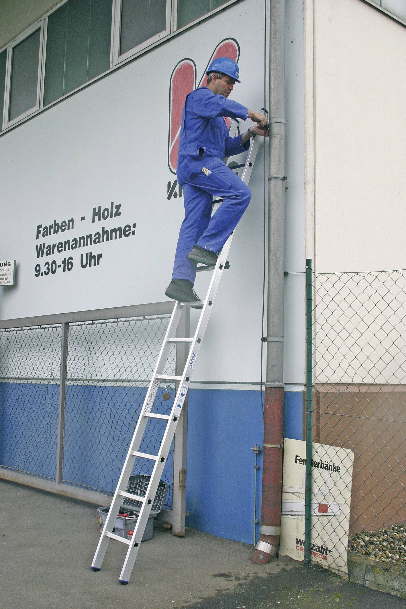 Une personne vêtue de vêtements de travail bleus est debout sur une échelle et travaille sur un mur de bâtiment à côté d'un panneau indiquant les heures d'ouverture.