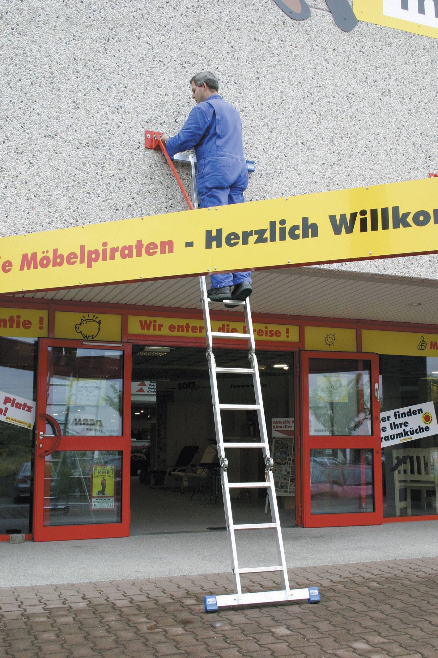 Un homme est debout sur une échelle et travaille sur la façade d'un magasin de meubles, sur laquelle est écrit : 'Bienvenue'.
