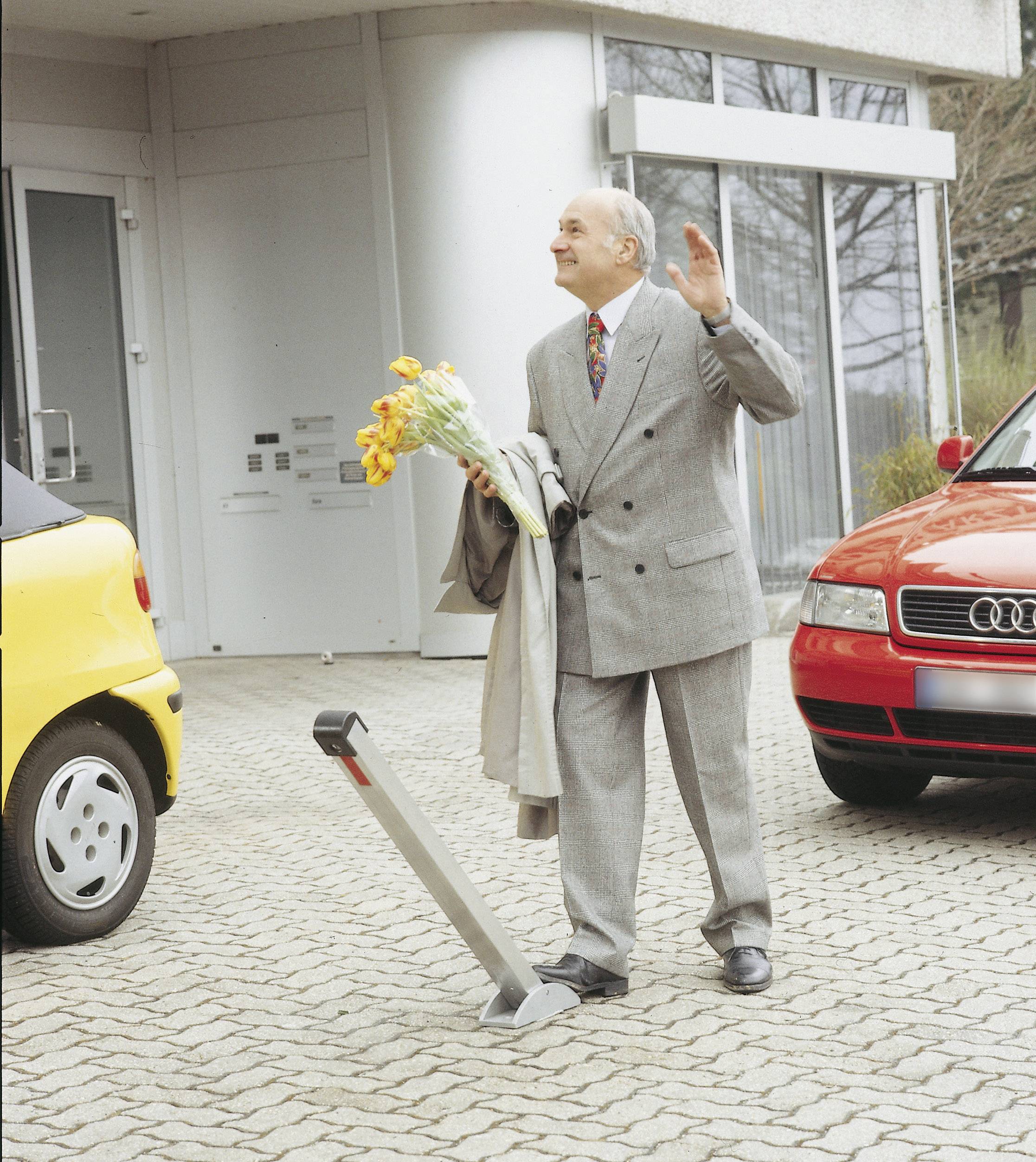 Un homme âgé en costume tient un bouquet de fleurs et fait signe. Il se trouve devant un bâtiment, entouré de voitures garées.