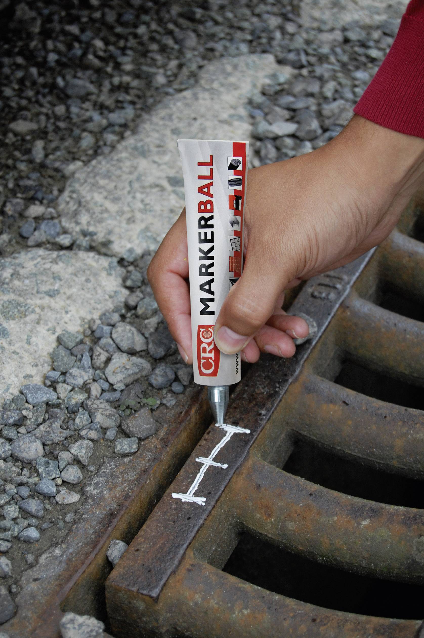 Une main dessine une ligne blanche sur une grille métallique avec un marqueur pour indiquer la présence de rouille. Des graviers sont visibles en arrière-plan.