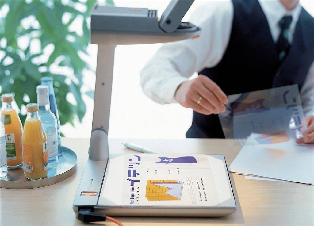 Un homme présente des diagrammes sur un rétroprojecteur posé sur une table. Des boissons sont disposées en arrière-plan.