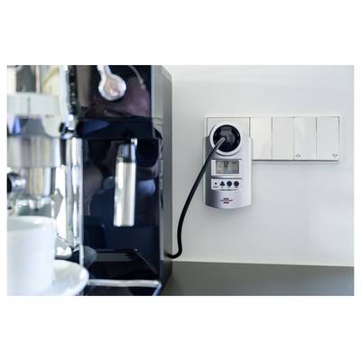 Električni aparat za kavu s utičnom mjernom jedinicom za potrošnju energije, pored zidnih prekidača u kuhinjskom ili kafićskom okruženju.
