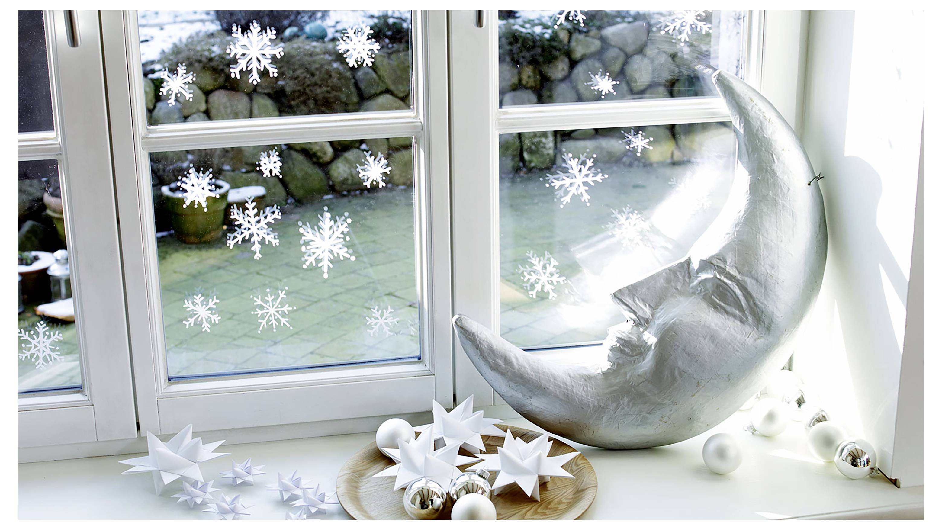 Dekorativni zimski izlog na prozoru sa viseći pahuljama, papirnatim zvijezdama i srebrnom skulpturom mjeseca na prozorskoj dasci.