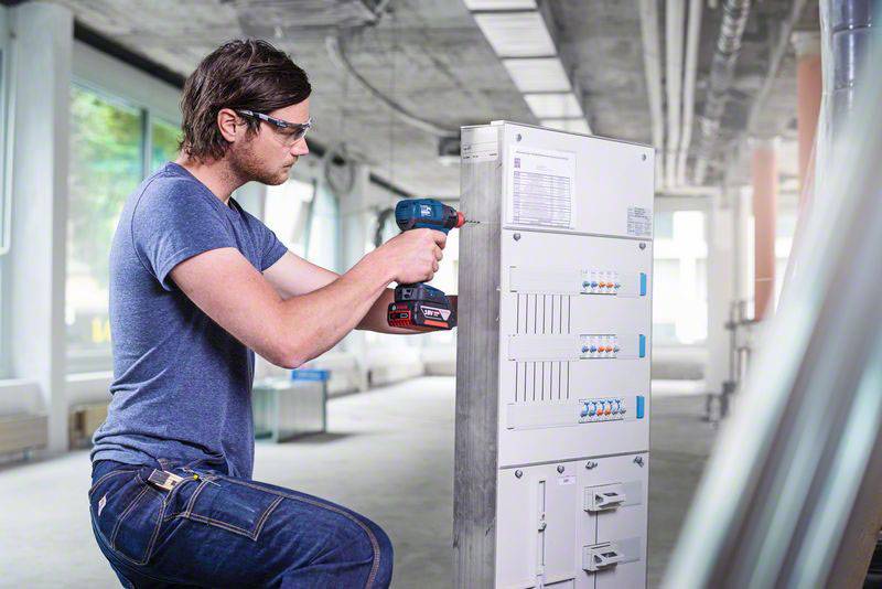 Muškarac u radnoj odjeći i zaštitnim naočalama koristi bušilicu kako bi radio na elektro ormaru u industrijskom prostoru.