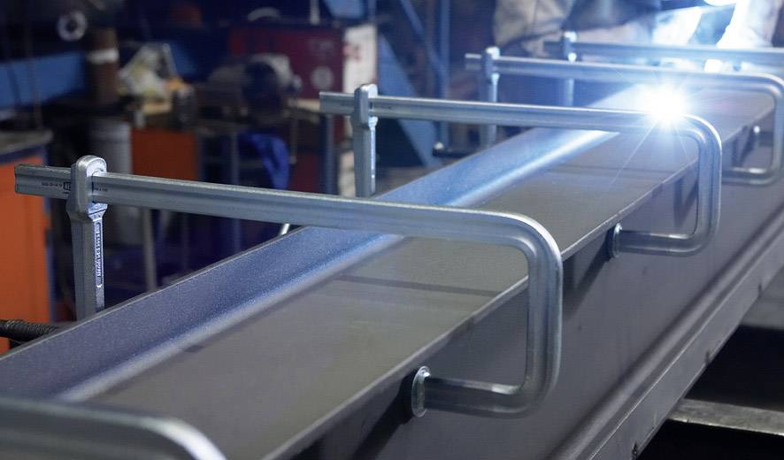 Metalac zavaruje veliki metalni nosač u radionici, okružen alatima i strojevima.