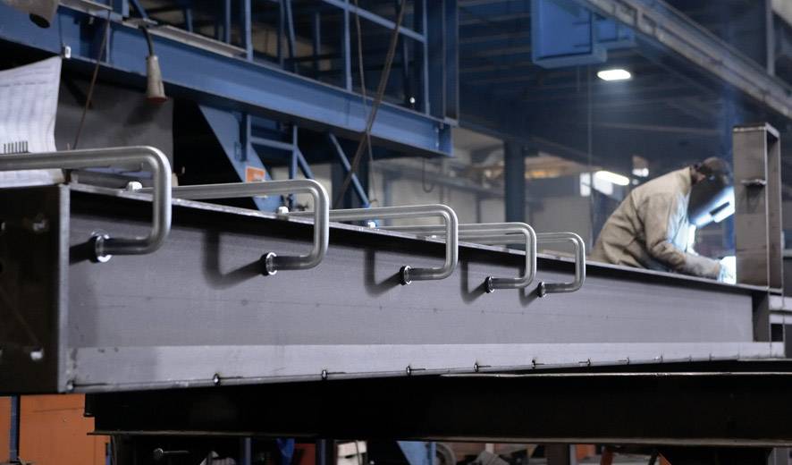 Vars zavarač radi na velikom metalnom nosču u industrijskoj hali. U prvom planu su cijevi pričvršćene za nosač.