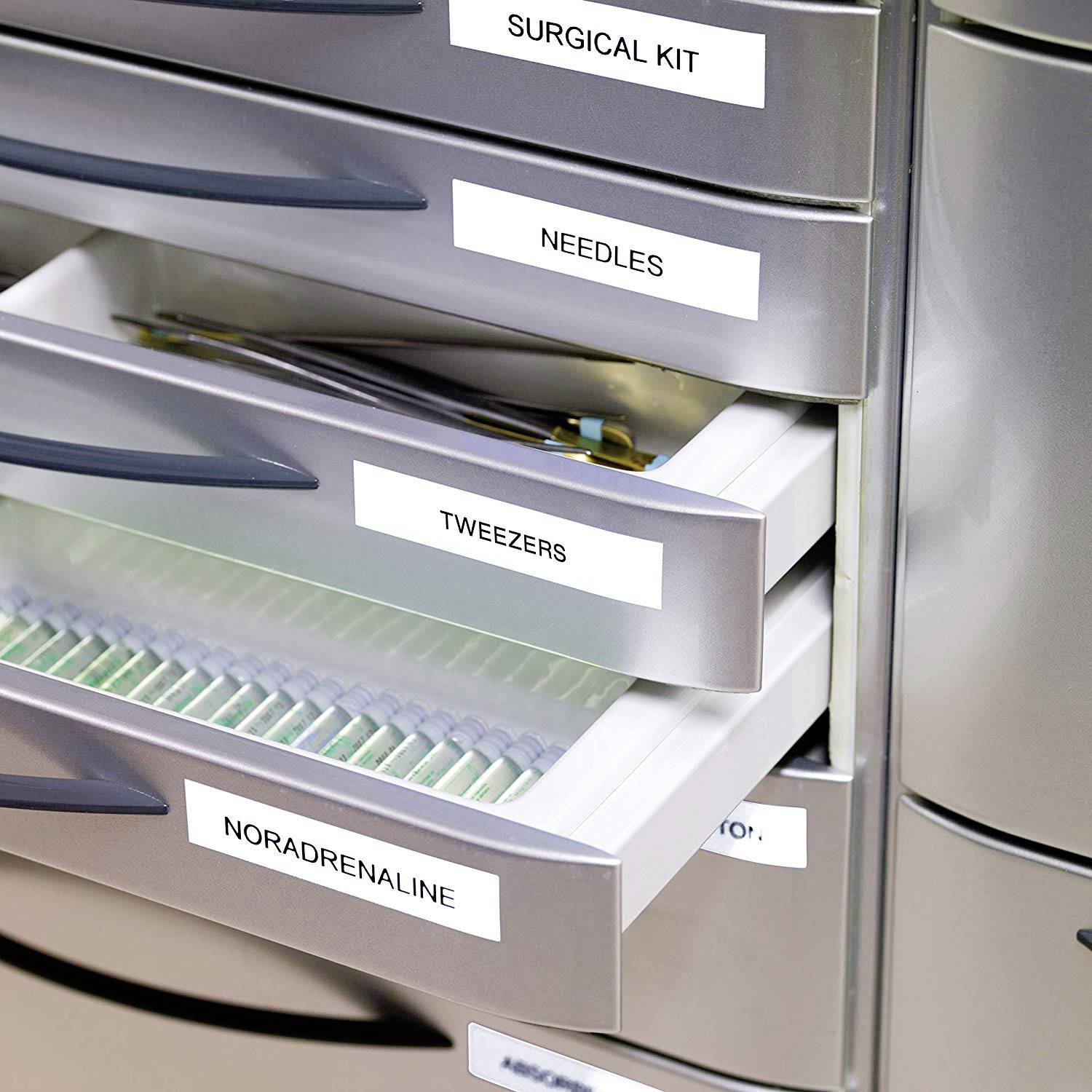 Otvoreni metalni ormar u medicinskom okruženju prikazuje precizno označene ladice, uključujući 'Noradrenalin' i 'Pincete'.