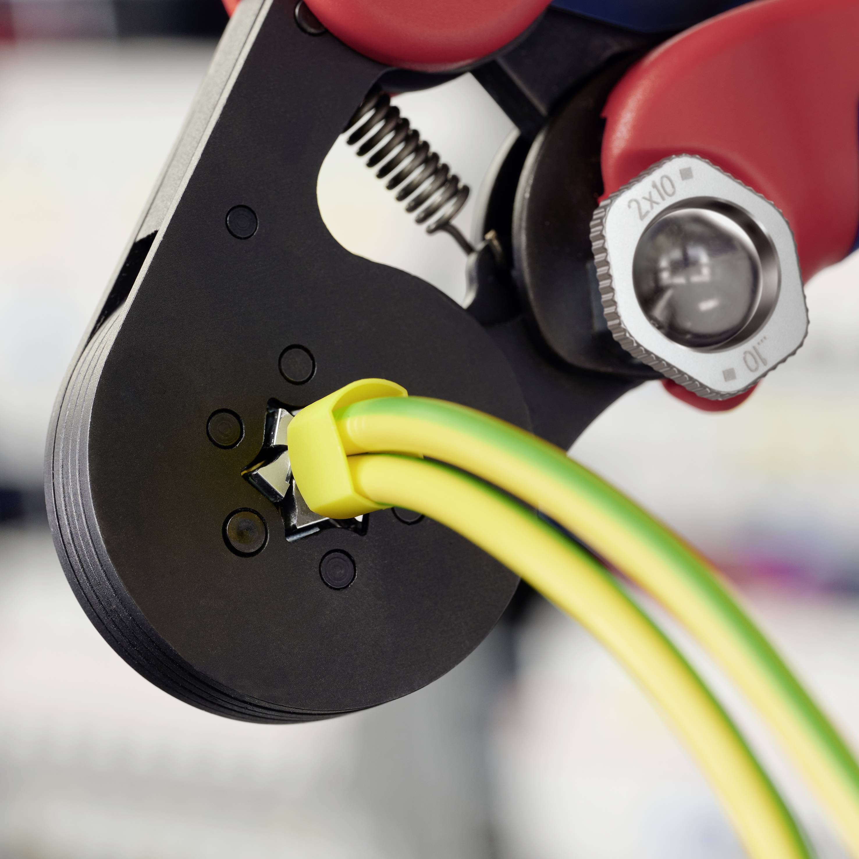 Kliješta koja krimpu žuto-zeleni električni kabel, približni kadar. Šarniri alata i vijak vidljivi, pozadina zamućena.