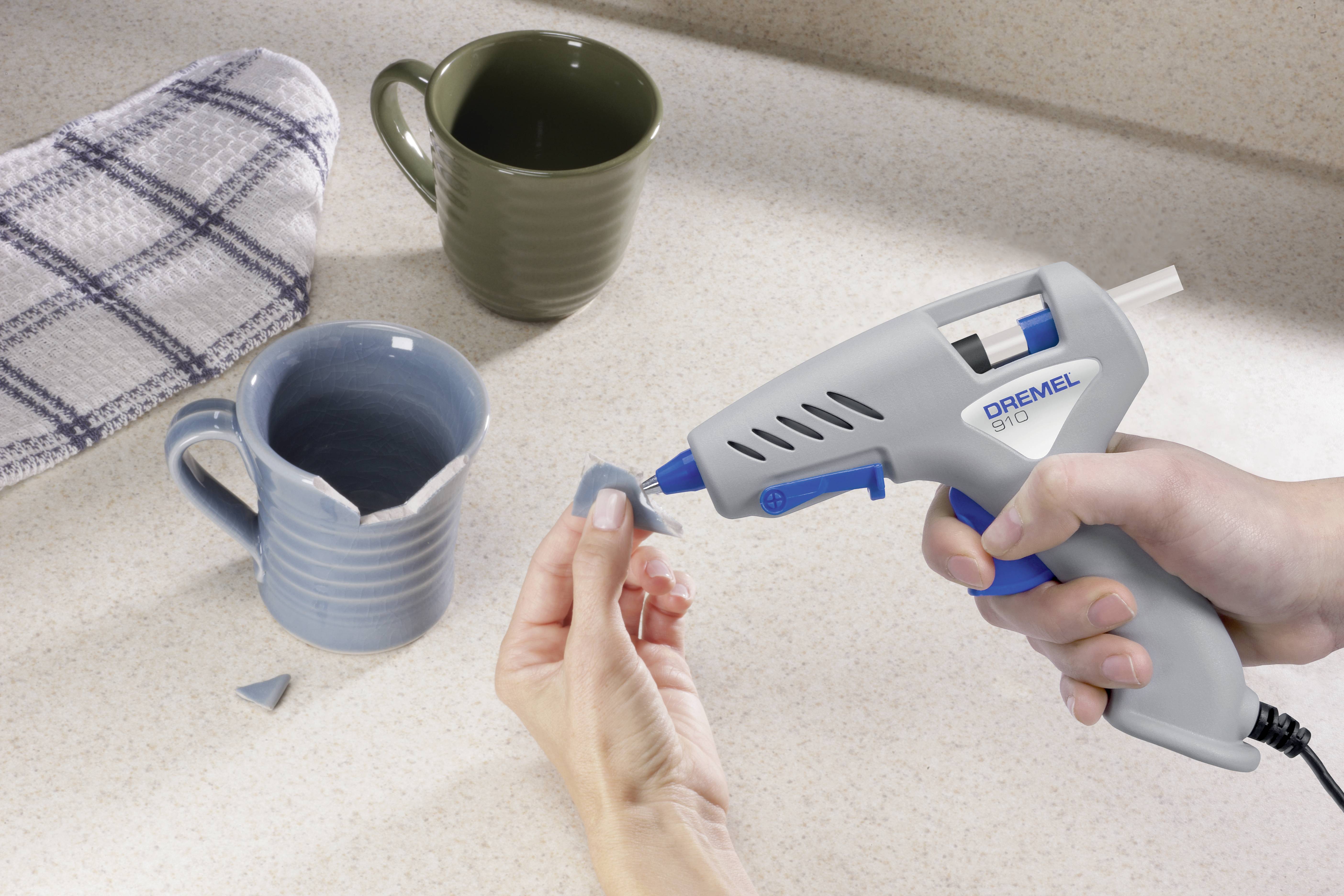 Osoba popravlja slomljenu plavu šalicu vrućom pištoljom za ljepljenje na kuhinjskoj radnoj ploči. U pozadini su dvije šalice.