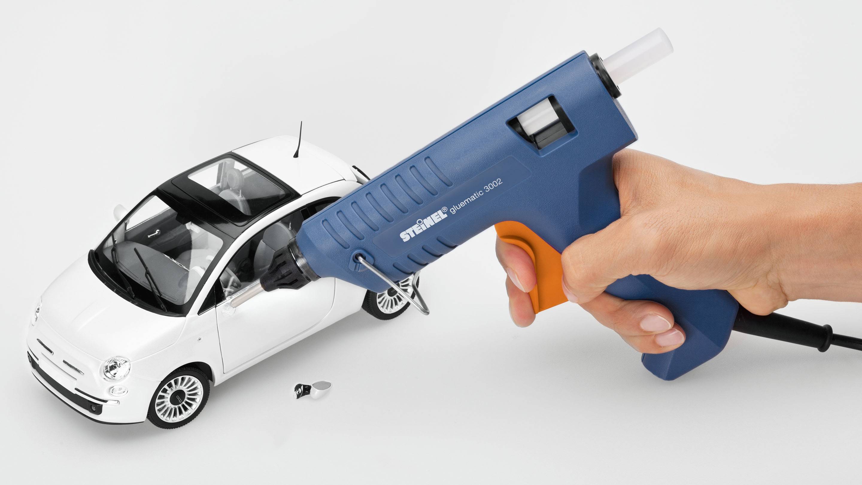 Netko drži plavu pištolj za vruće ljepljenje blizu bijelog modela automobila, kao da ga popravlja.