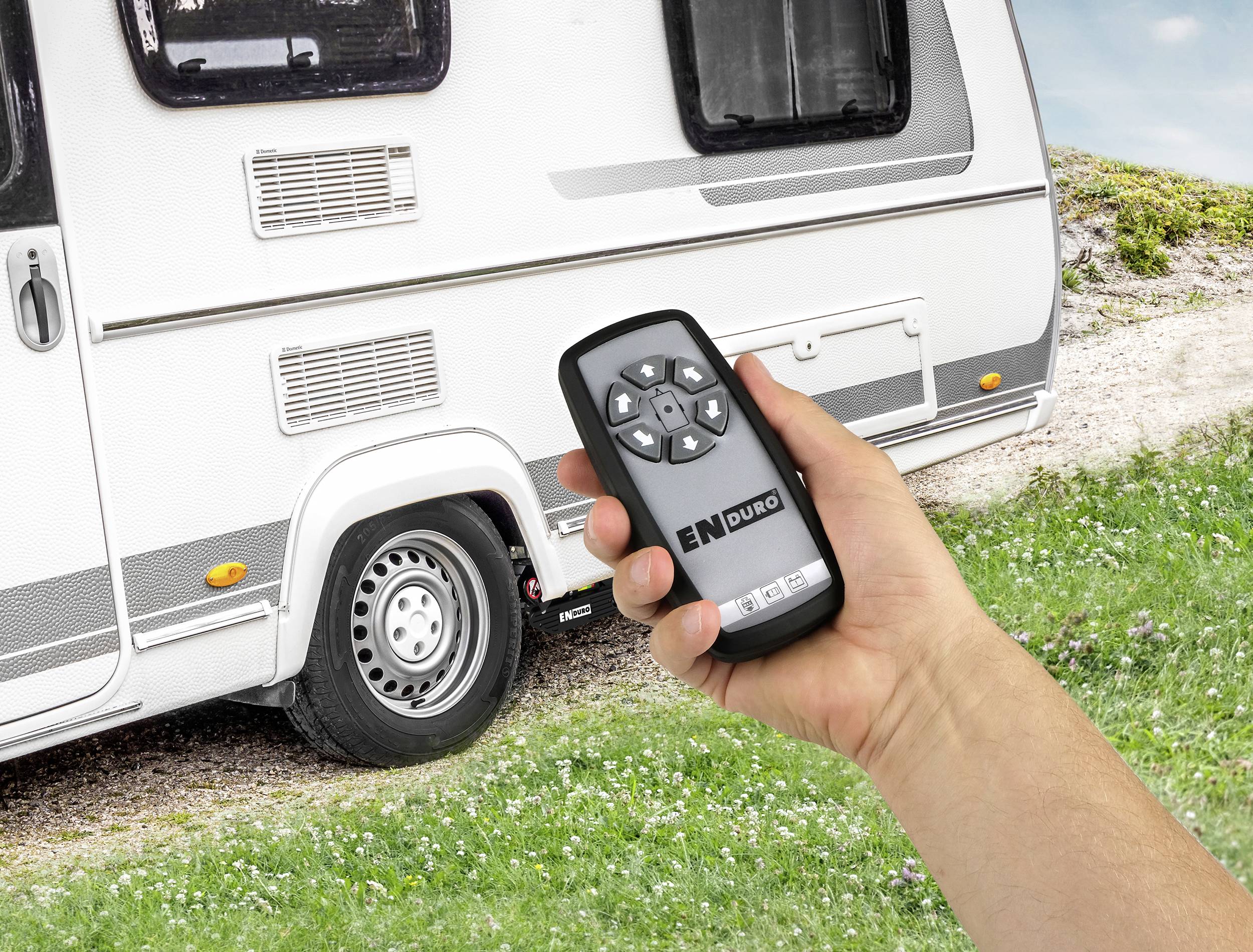 Približni kadar ruke koja drži daljinski upravljač sa strelicama za smjer, u pozadini parkiran kamper na livadi.