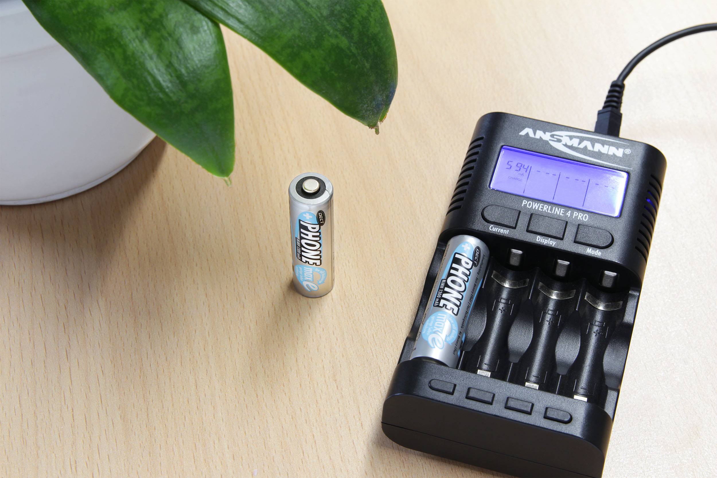 Punjač za baterije punjač četiri AA baterije na drvenom stolu. Jedna baterija stoji pored njega. Gore lijevo je vidljiv dio zelene biljke.