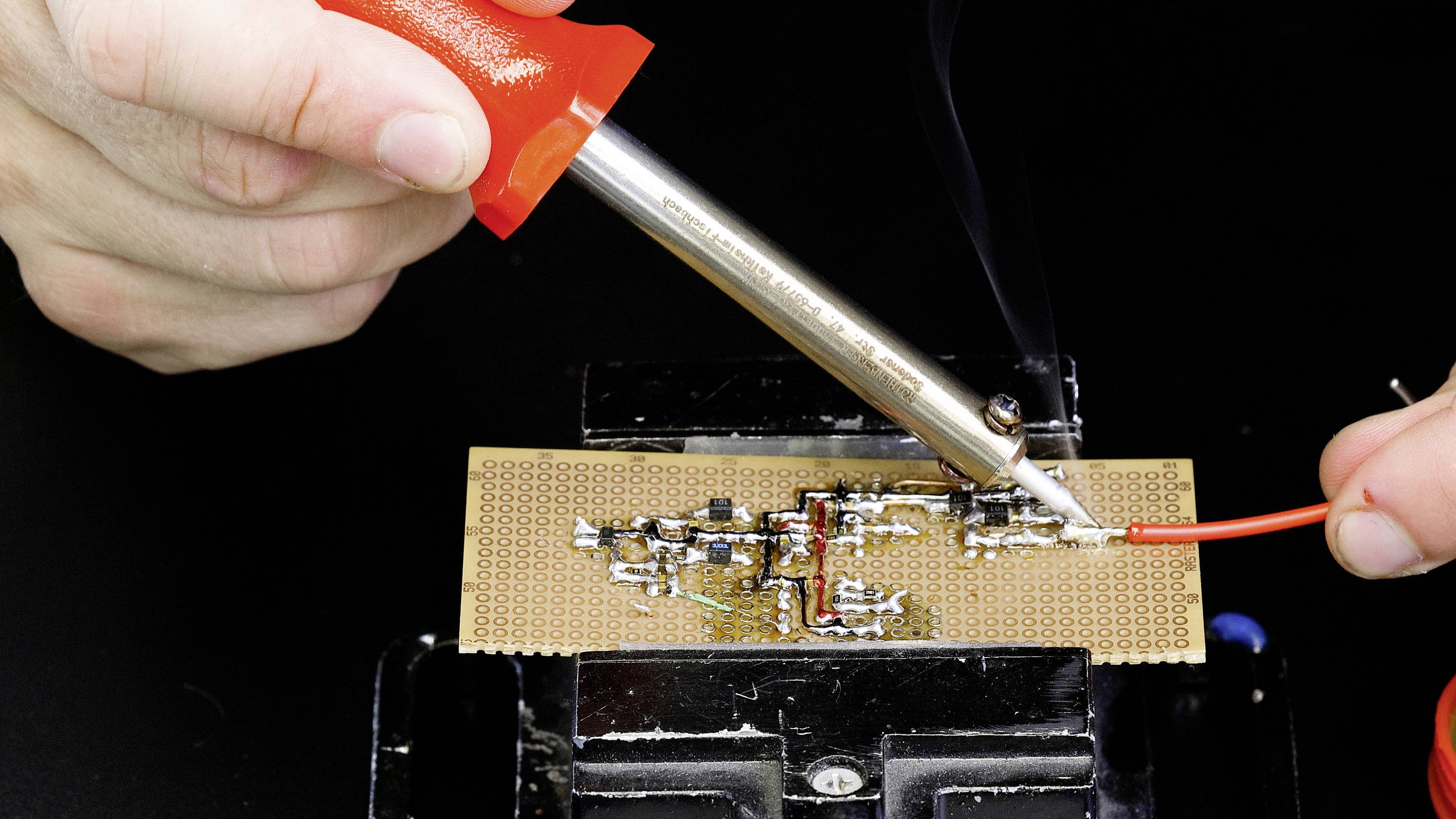 Jedna osoba lemom spaja crveni kabel na tisknoj pločici. Izrada ili popravak elektroničkih sklopova.
