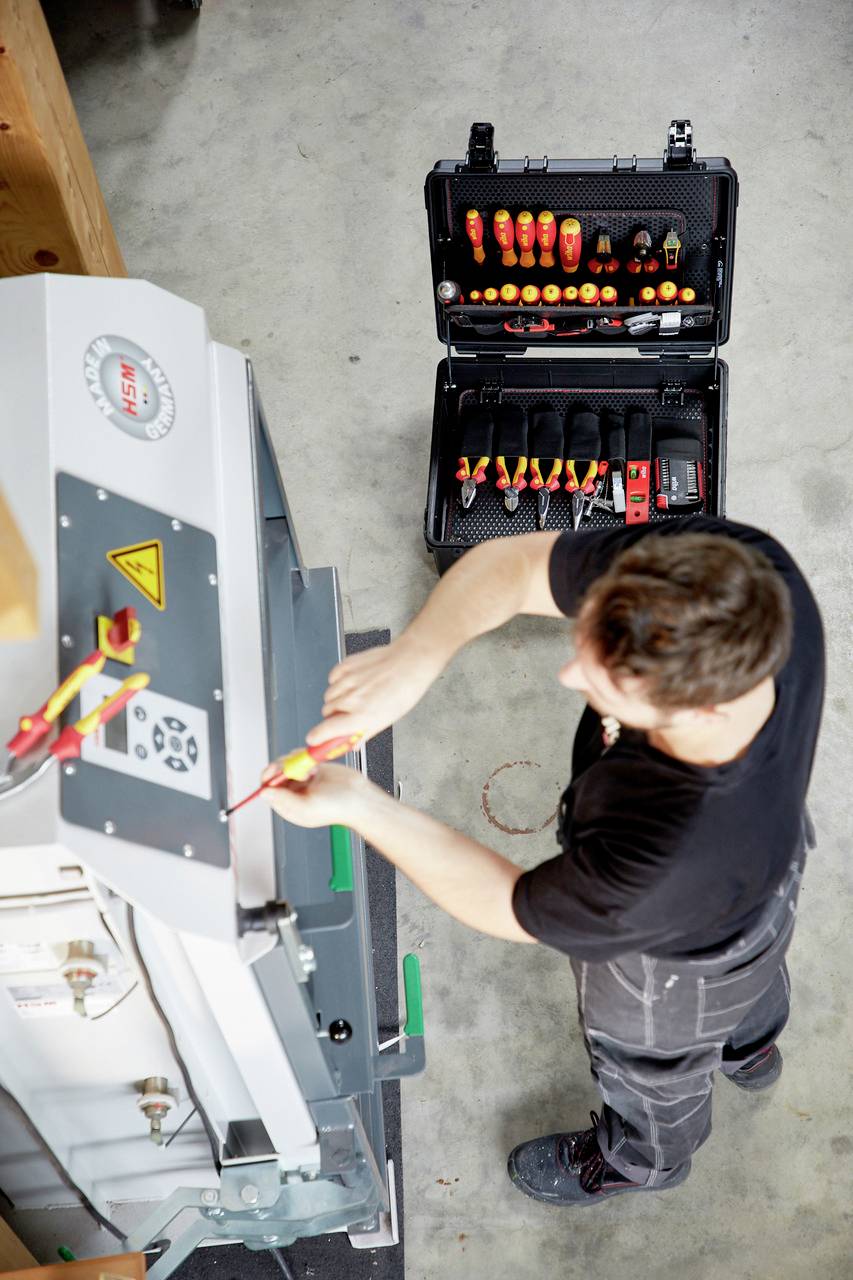 Jedna osoba popravlja stroj koristeći alate iz otvorenog alata. Naglasak je na preciznom radu i sigurnosnim upozorenjima vezanim za električnu opremu.