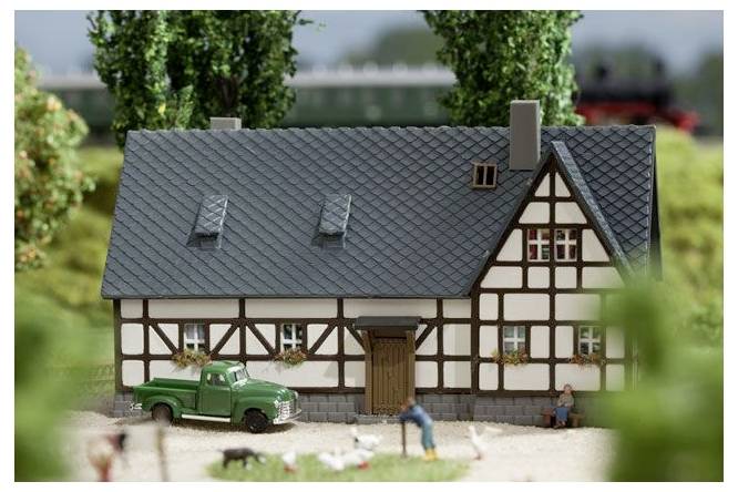 Minijaturni model半-drvene kuće sa zelenim pick-up kamionom i sitnim figuricama ispred, smešten u uređenu krajoliku.