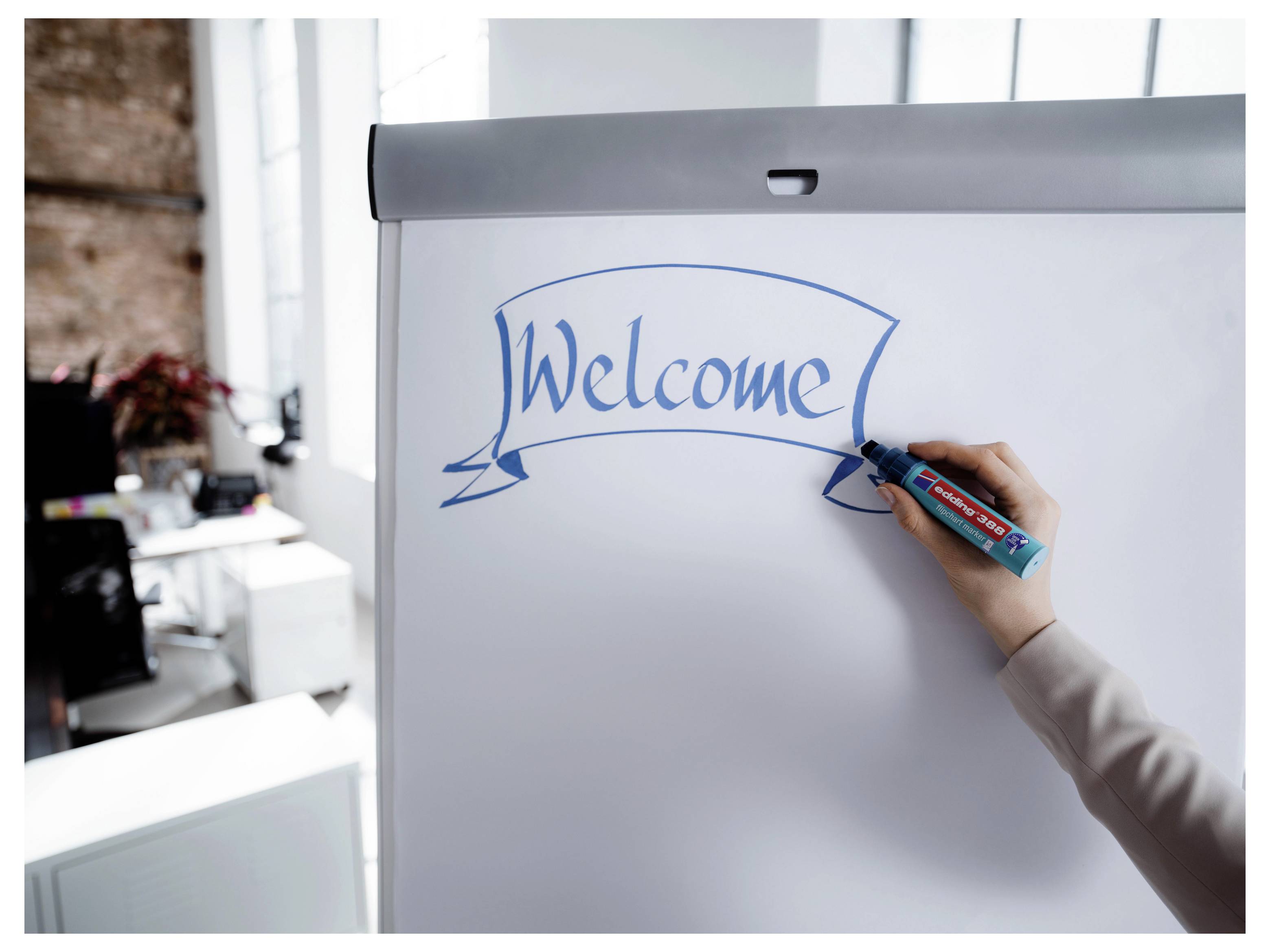 Ruka piše 'Welcome' (Dobrodošli) u plavoj boji na flipchartu u uredskom okruženju.