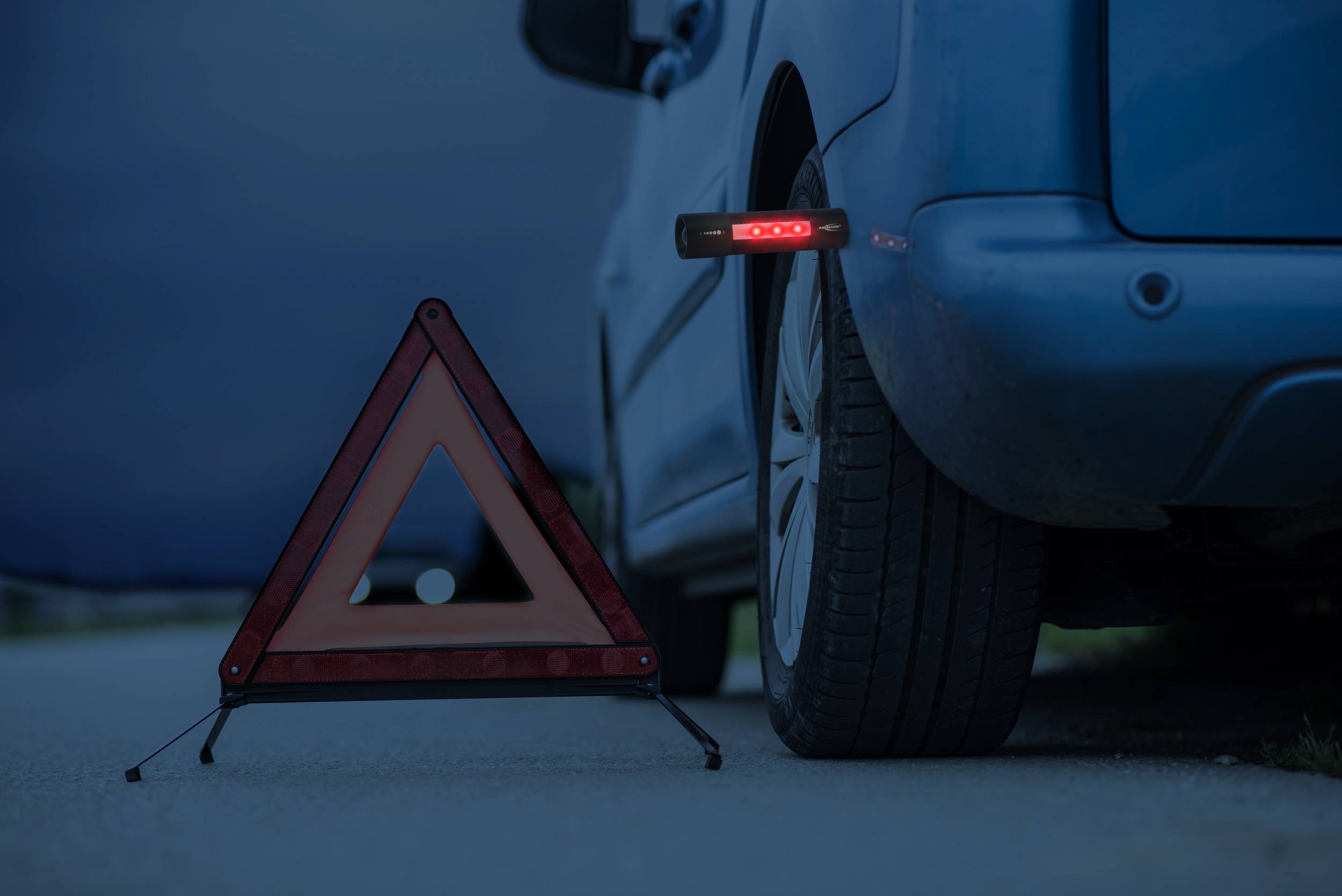 Plavi automobil s probitom gumom stoji uz rub ceste. Sigurnosni trokut s reflektirajućim rubovima postavljen je ispred automobila.