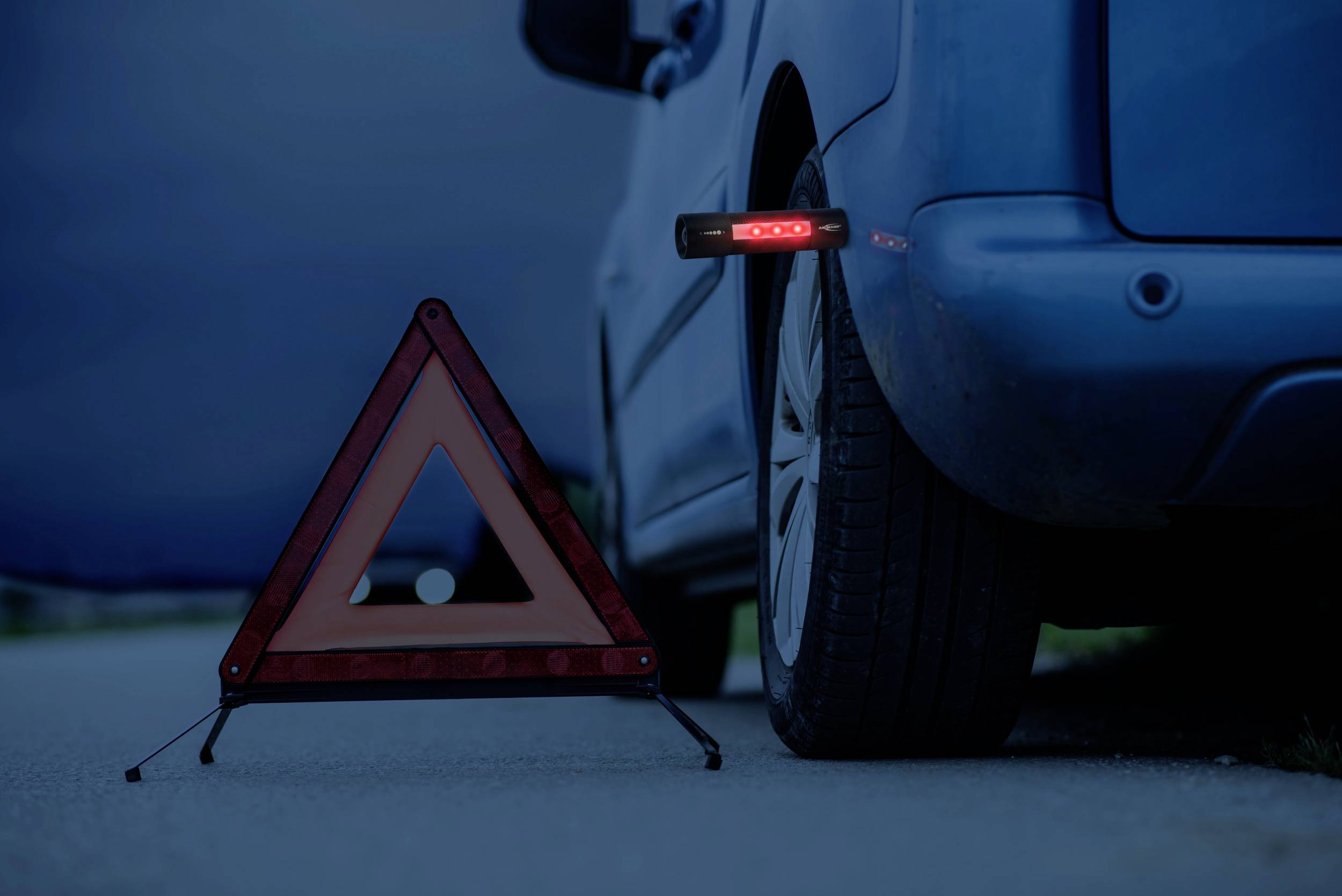 Žuto trepereće sigurnosno trokutasto znanje stoji blizu automobila parkiranog uz rub ceste u sumrak, upozoravajući na moguću opasnost.