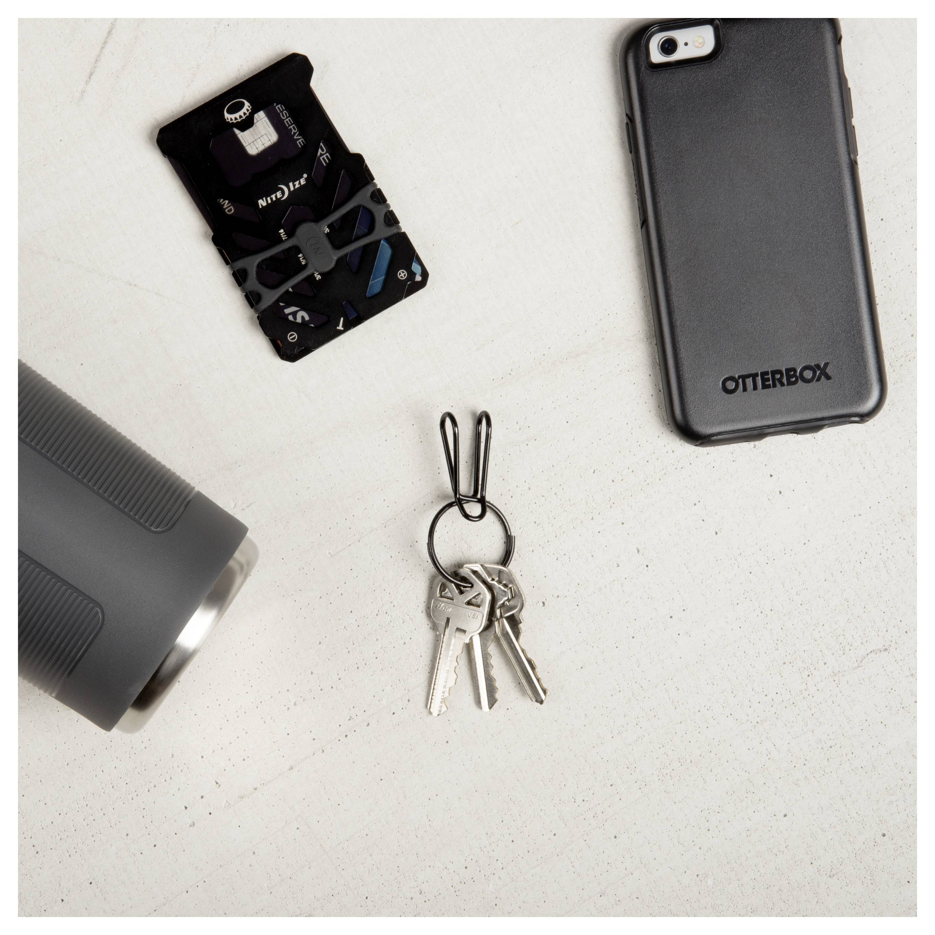 Ravni prikaz (flat lay) silikonskog zaštitnika za smartphone, multifunkcijske kartice, metalnog ruba čaše, i ključeva s crnim nosačem na svijetloj podlozi.
