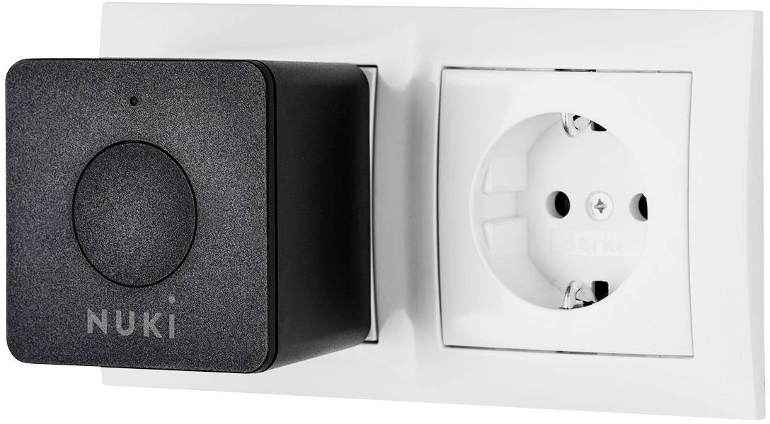 Crni Nuki Smart Plug postavljen je pored bijele zidne utičnice. Smart Plug proširuje utičnicu pametnim funkcijama.