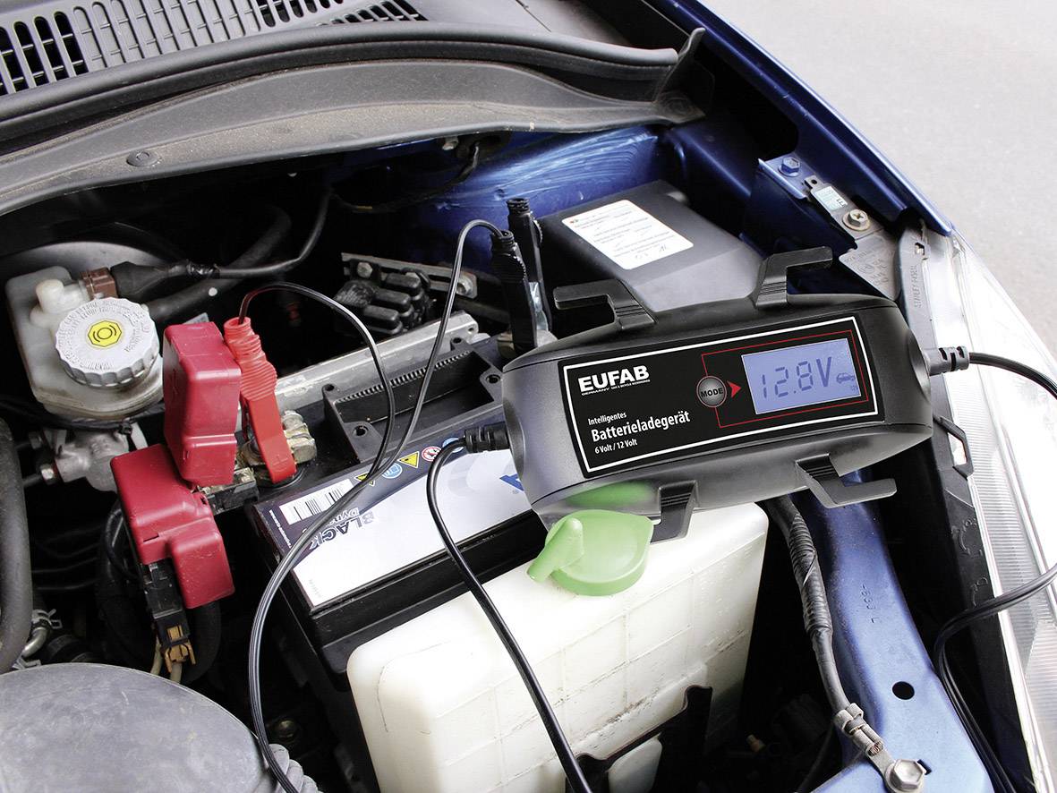 Punjač baterije s digitalnim zaslonom pokazuje 12,8V, priključen na autobateriju u prostoru motora vozila.