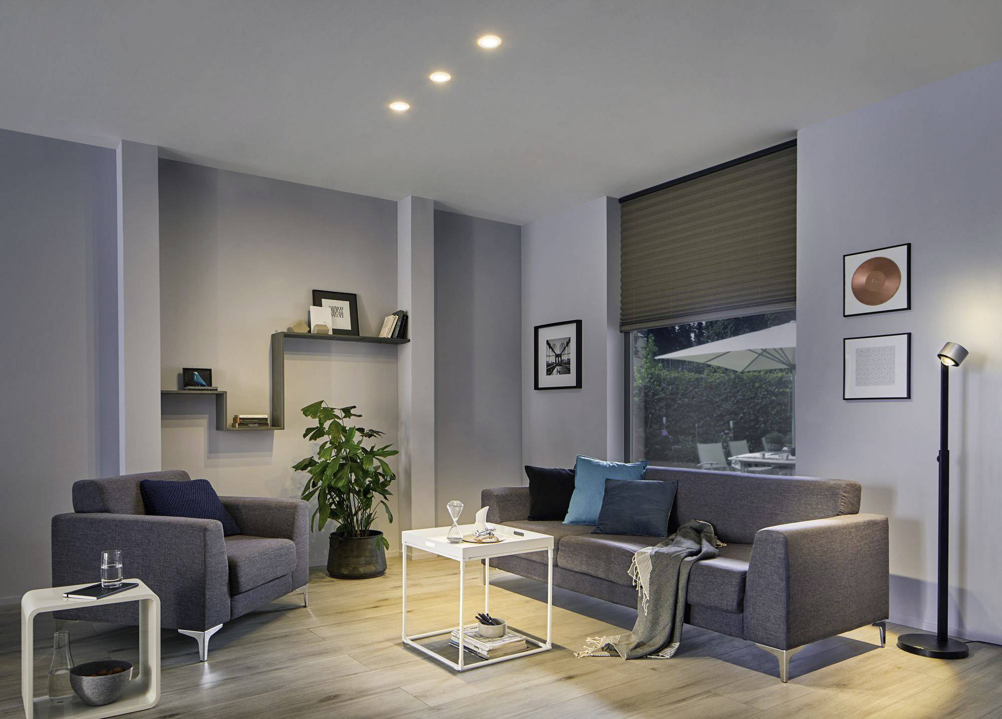 Dnevni boravak sa sivom kaučem, foteljom, stolom za kavu i podnom lampom. Na zidu se nalaze police sa dekoracijom. Prozor s pogledom van.