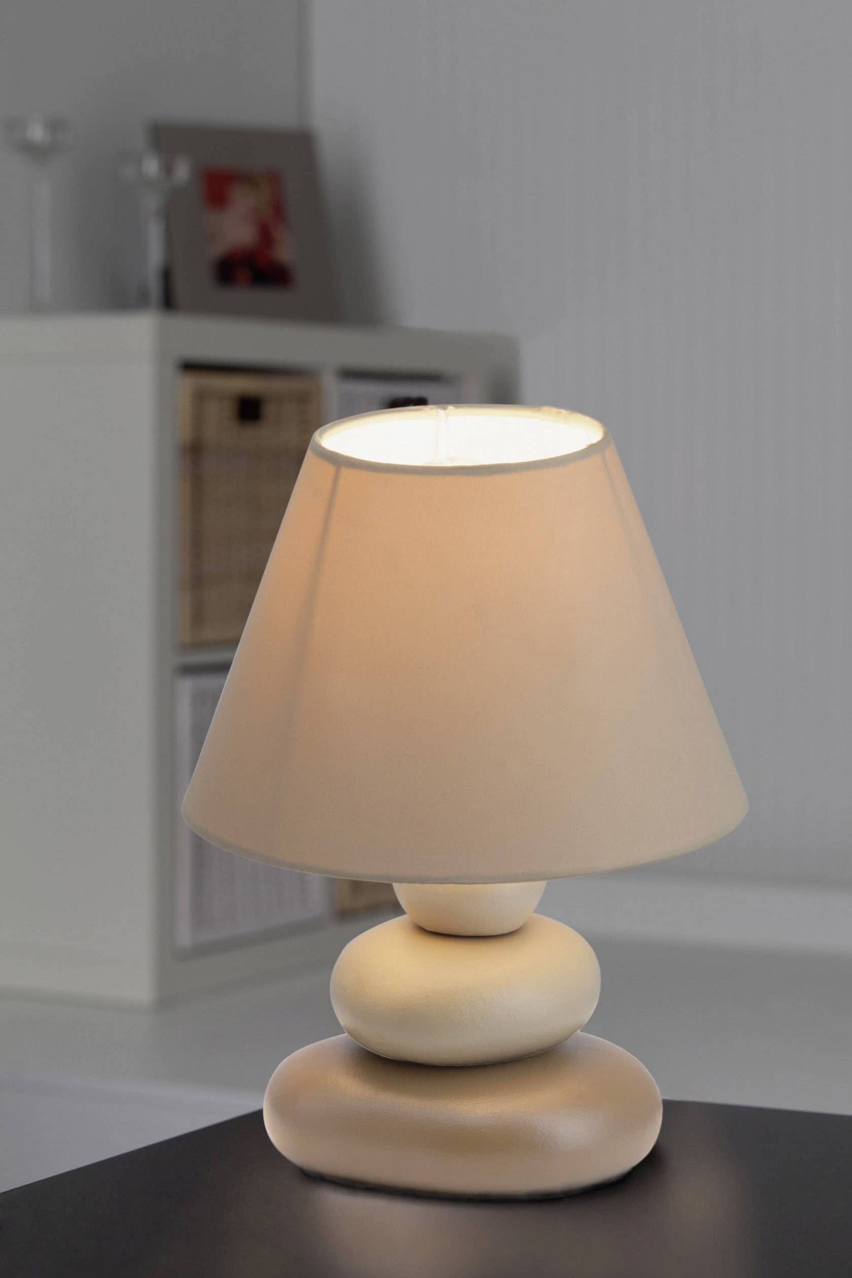 Mala stolna lampa sa stopaljem u obliku kamena i bež sjenilom stoji na tamnom stolu, a u pozadini se nalazi polica s košarama.