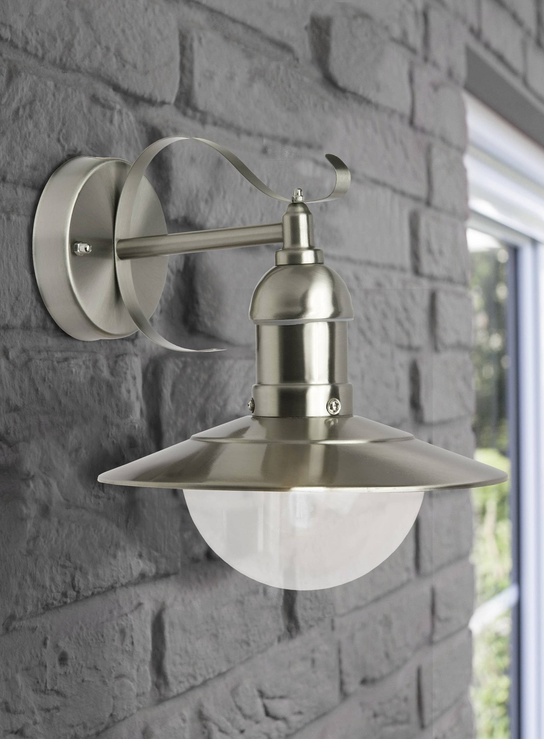 Moderna zidna lampa od brušenog metala visi na opečnatom zidu, pored prozora kroz koji se nazire zeleno raslinje.