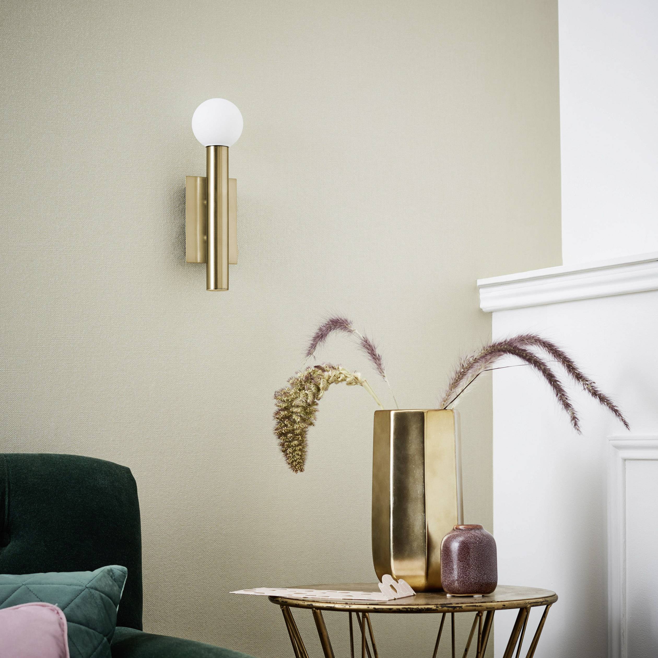 Zlatna zidna lampa iznad zelene sofe u jednostavnom, modernom prostoru. Vaza sa sušenim cvijećem i mala skulptura na stolu.