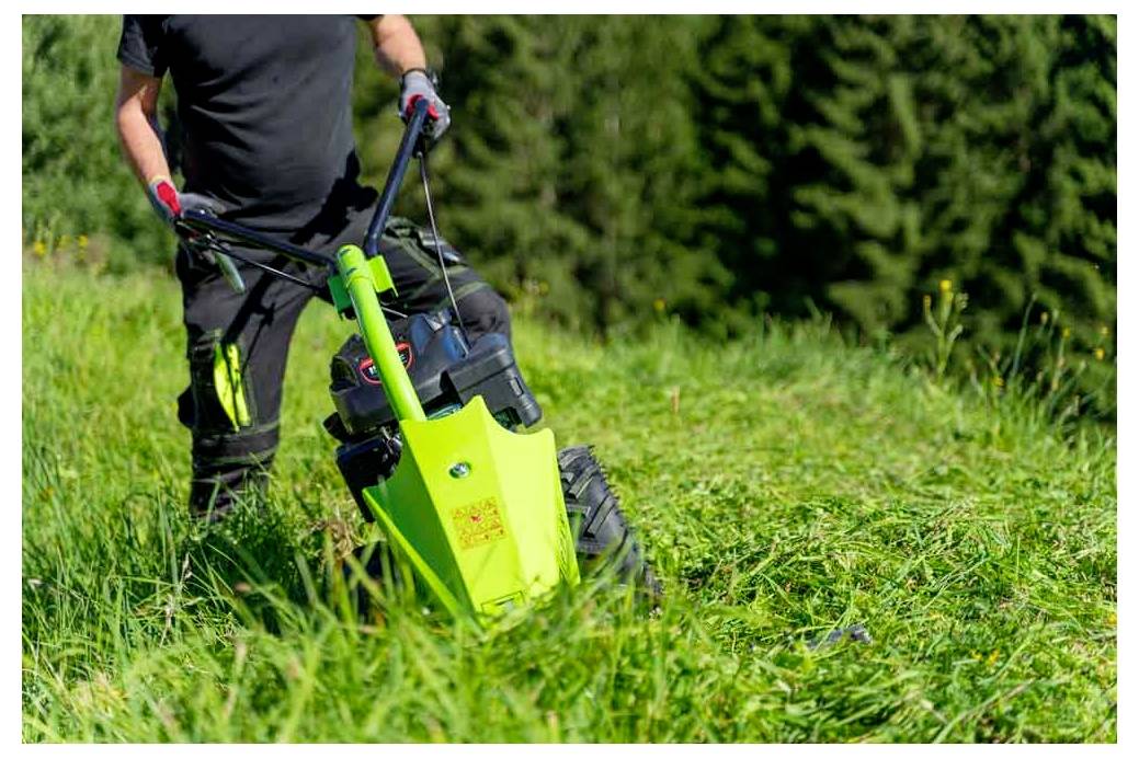 Osoba rukuje zelenom kosilico na travnatom polju, okružena drvećem. Radnja upućuje na vanjsko održavanje ili vrtlarenje u tijeku.