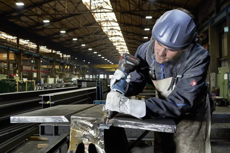 Radnik u zaštitnoj opremi koristi električni alat za rad na velikom komadu metala u industrijskom skladišnom prostoru.