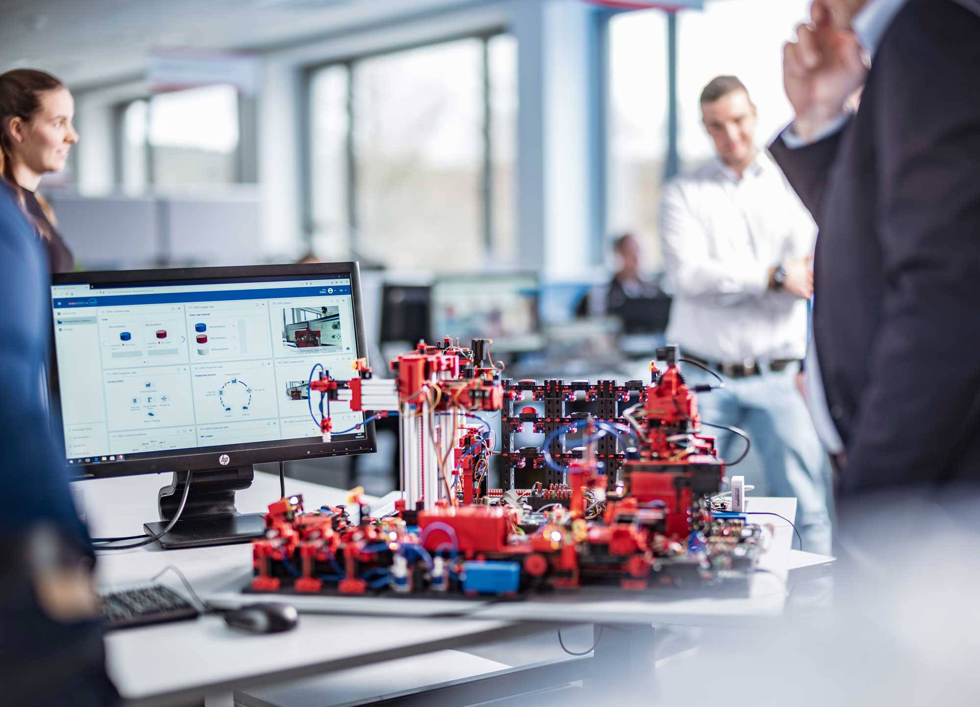 Grupa ljudi u uredu promatra kompleksan model na stolu. Monitor prikazuje tehničke dijagrame.