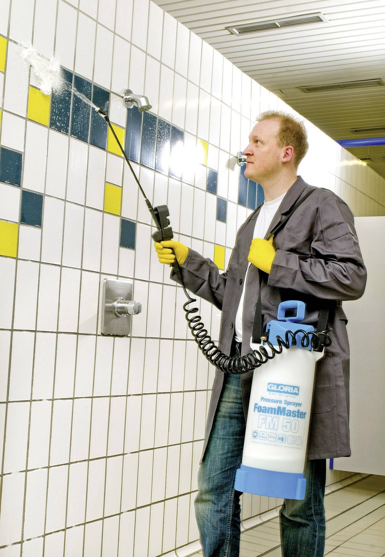 Muškarac čisti bijele pločice uređajem za pjenastu čišćenje. Žute i plave pločice čine uzorak na zidu.