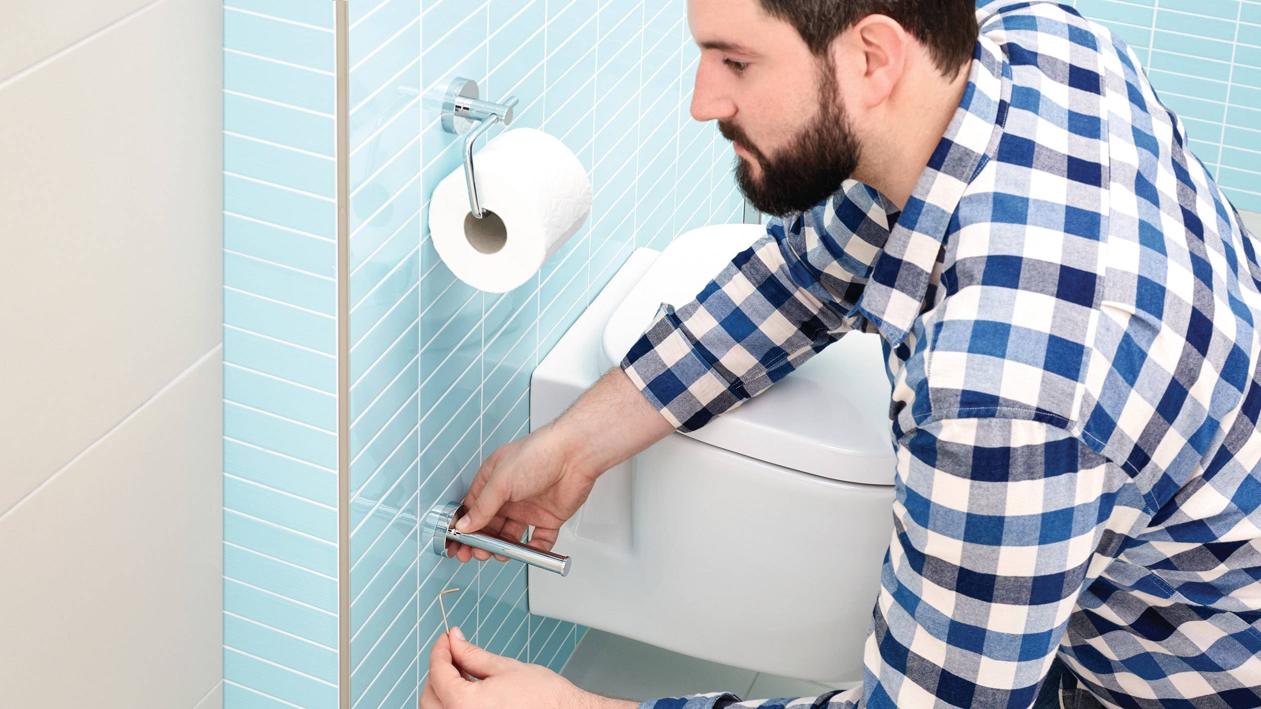 Muškarac u kariranoj košulji popravlja nosač role toaletnog papira u kupaonici s plavim pločicama.