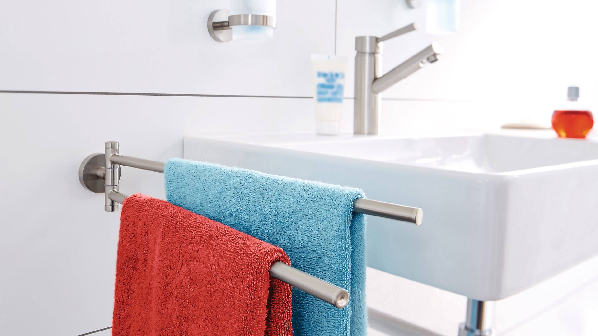Kupaonica s pravokutnim bijelim umivaonikom, dva zidna držača ručnika s crvenim i plavim ručnicima, jednom slavinom i kozmetičkim proizvodima.