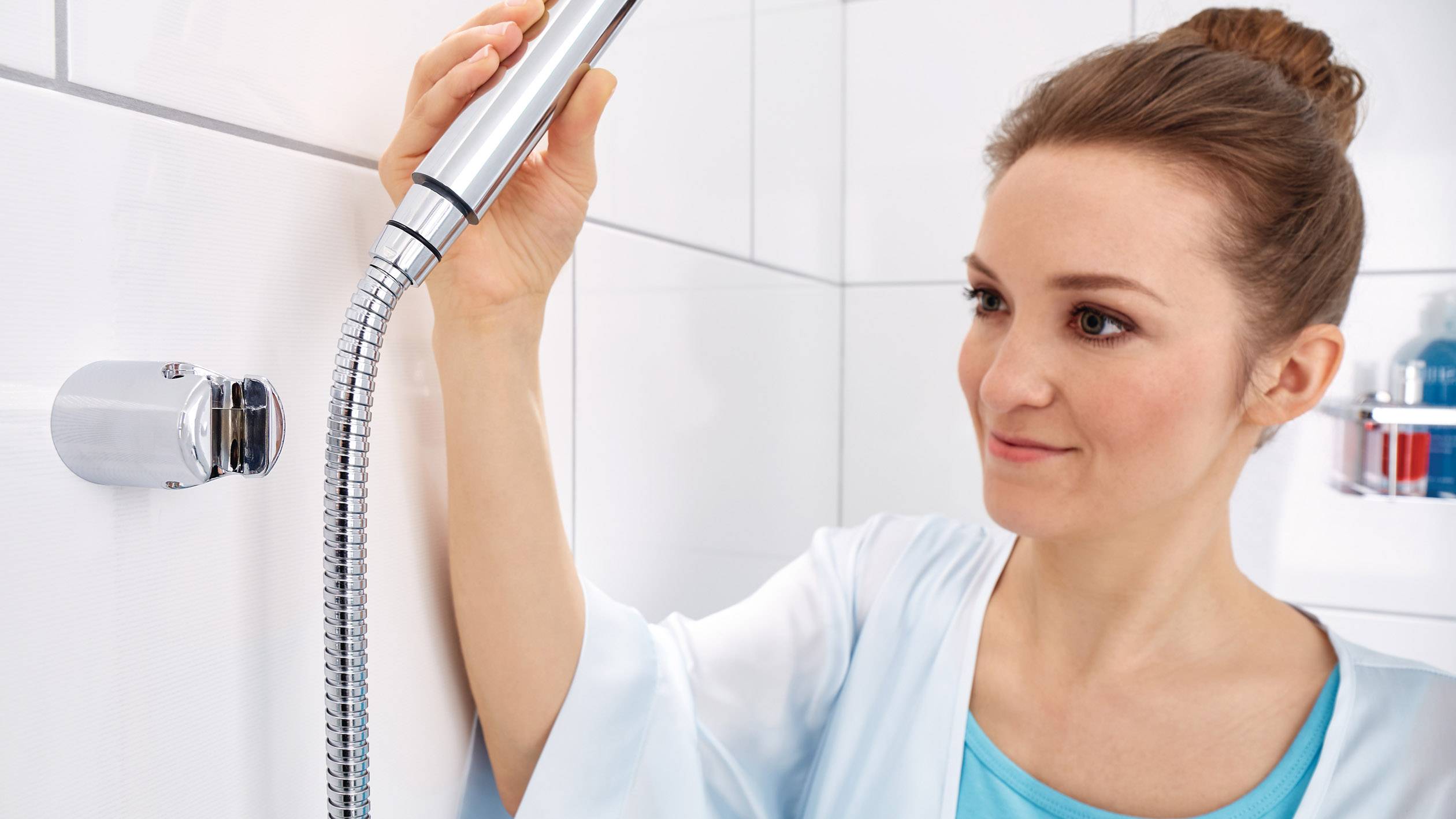 Žena podešava tuš glavu u svjetloj kupaoni. Nosi plavi ogrtač za kupanje i djeluje koncentrirano.