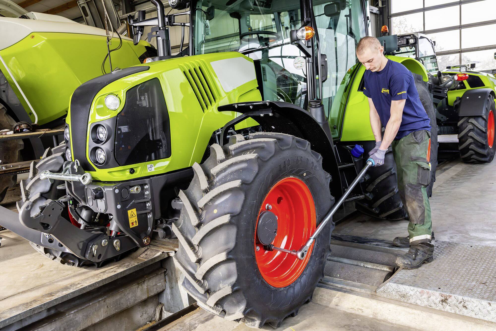 Mehaničar popravlja veliko poljoprivredno vozilo u radionici. Traktor je zelene boje s crvenim kotačem.