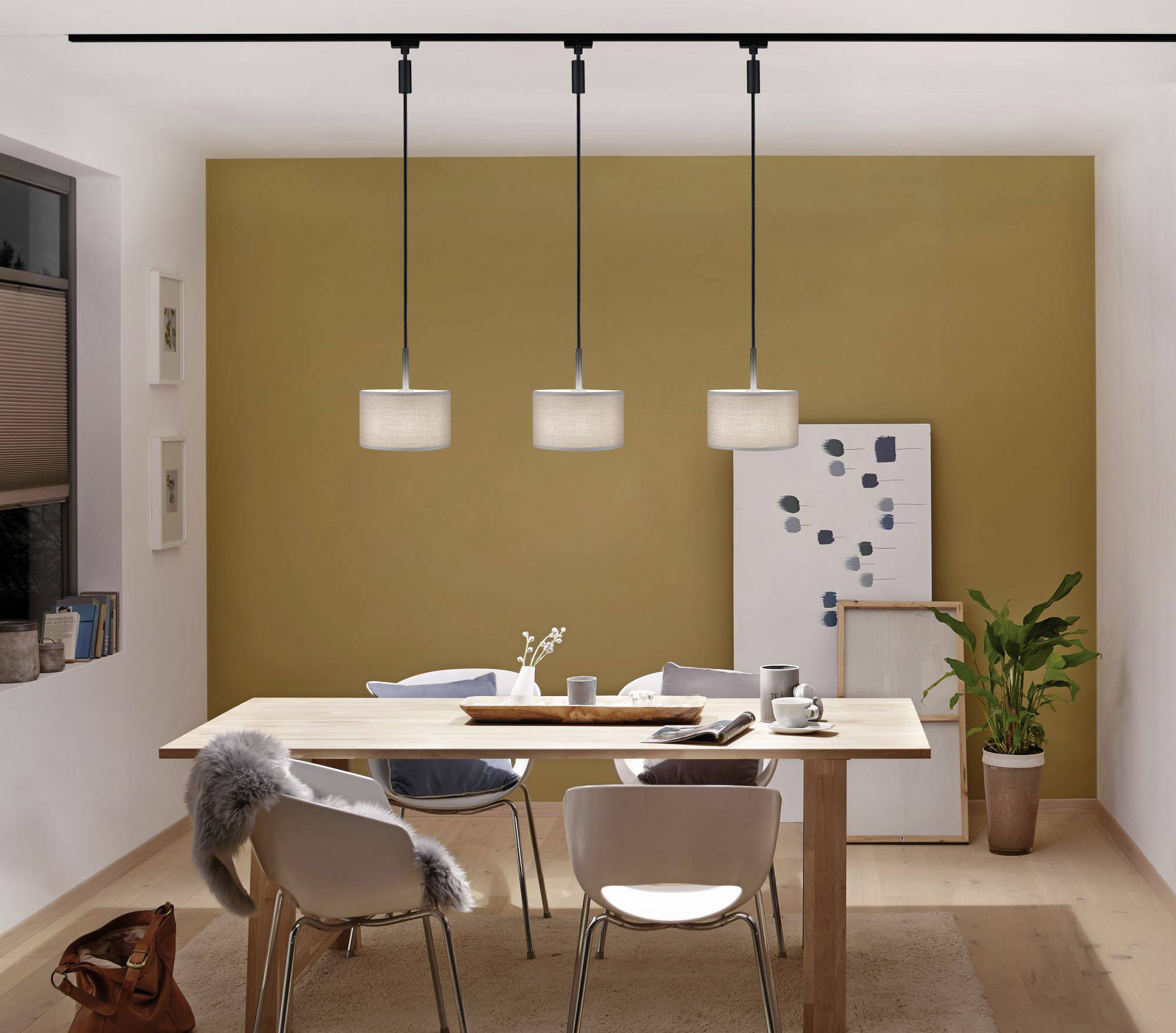 Blagovaonica s drvenim stolom i stolicama, bež zidovi i moderne stropne lampe. Bijela ploča za bilješke s notama i biljkom u kutu.