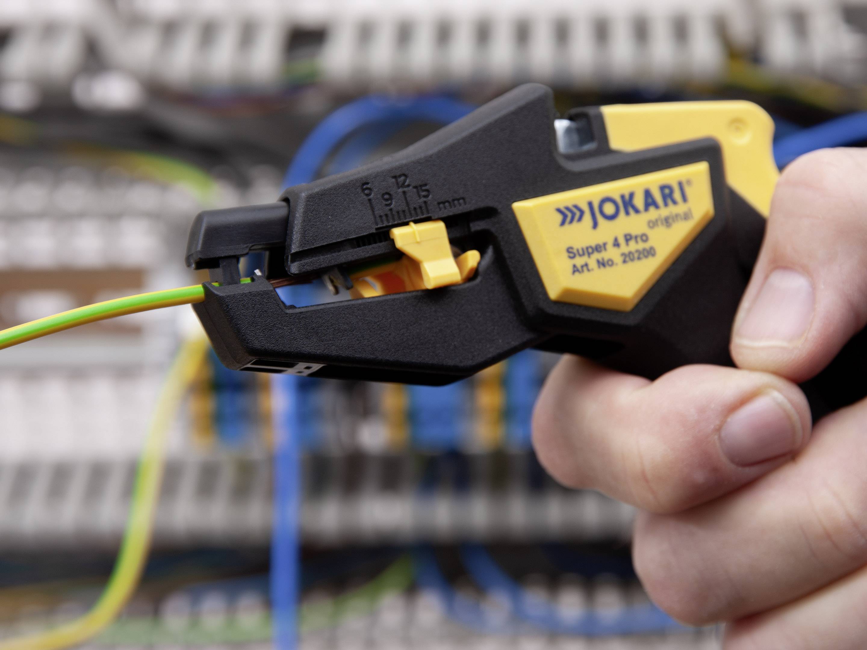 Jedna ruka drži skidač izolacije kako bi skinula izolaciju sa zeleno-žutog kabela. U pozadini su nejasno vidljive električne razvodne kutije.
