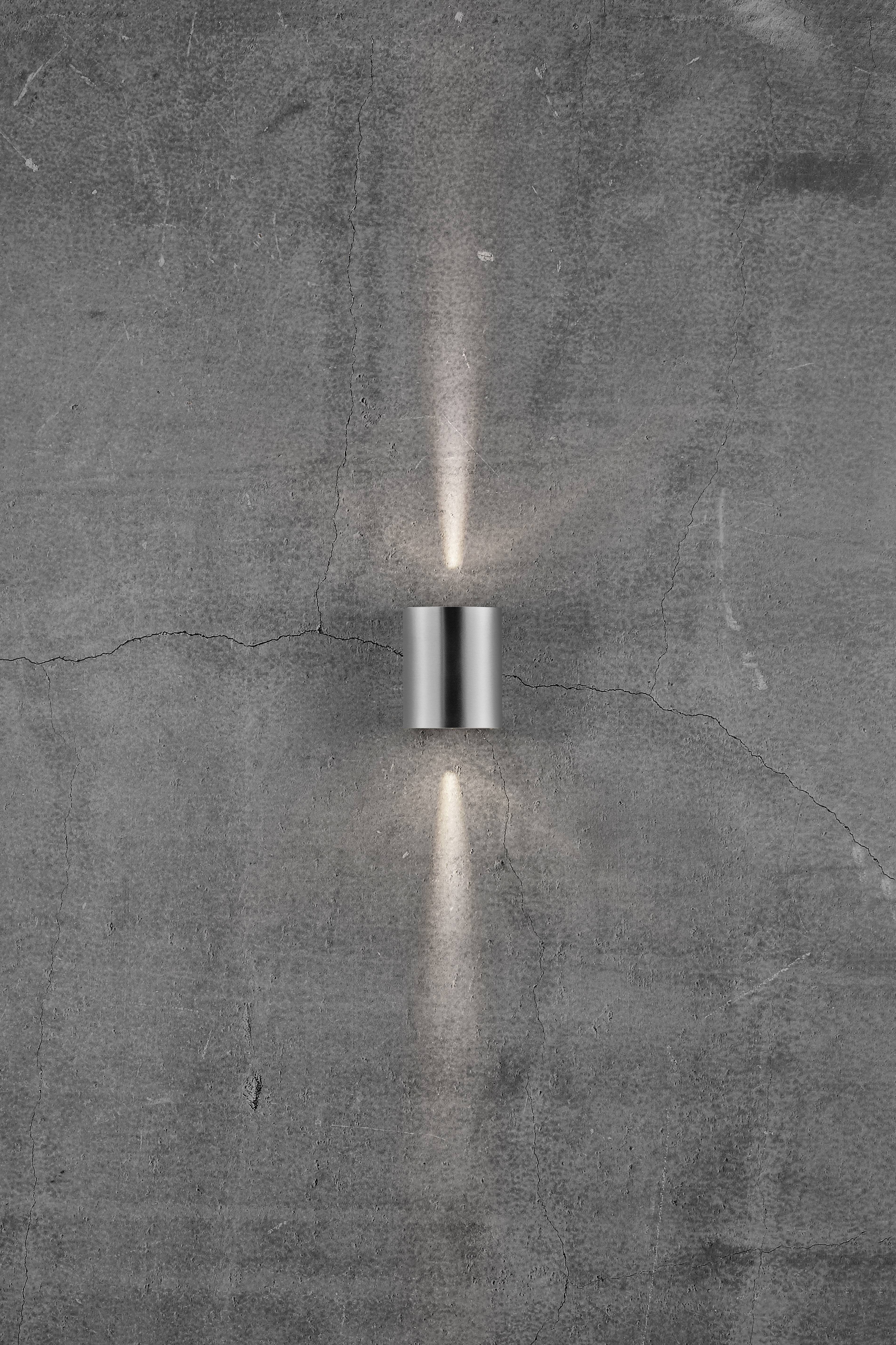 Lampa na sivom, strukturiranom zidu baca svjetlost gore i dolje. Dizajn djeluje moderan i minimalistički.