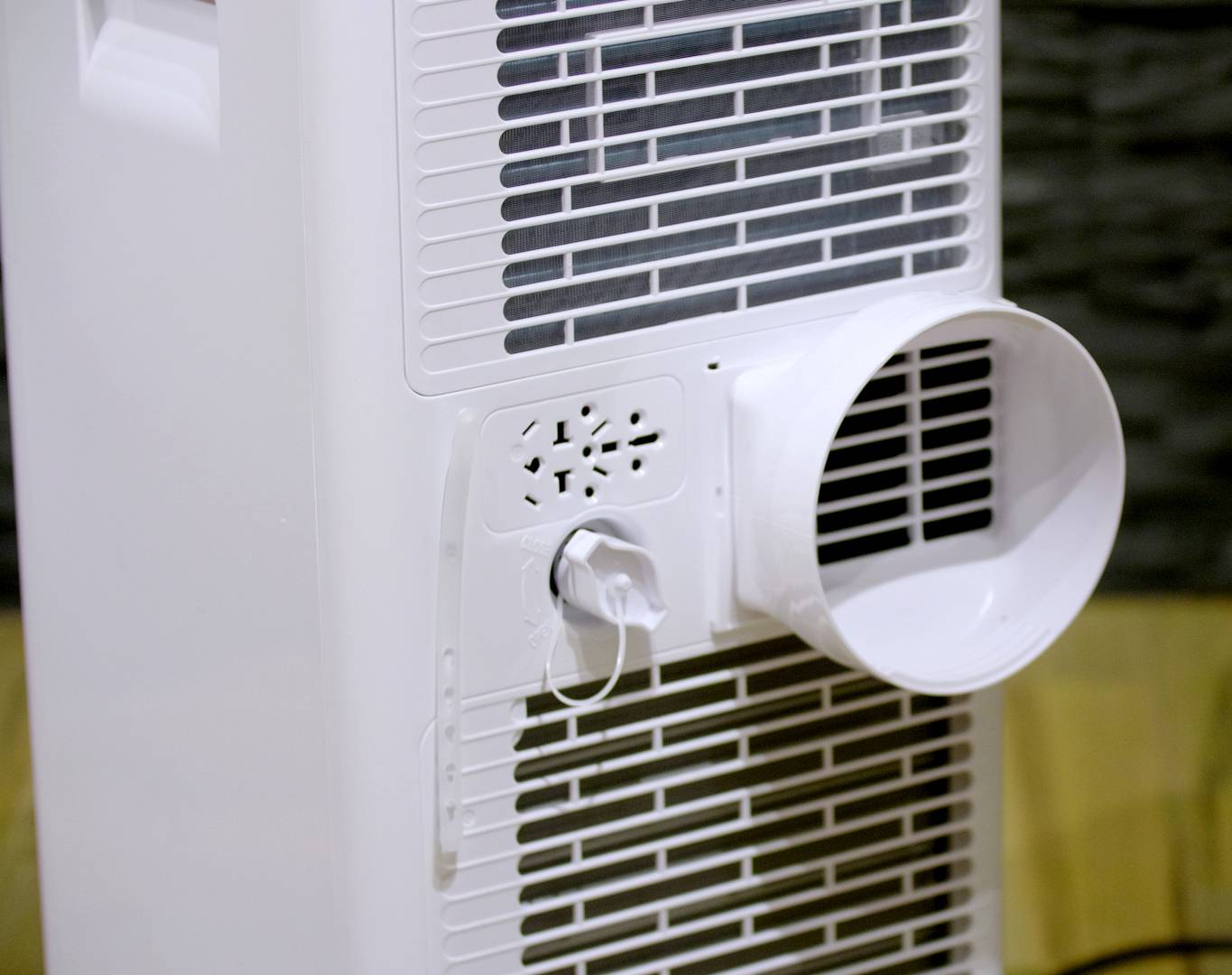 Bijeli pročistač zraka s ventilacijskom rešetkom i velikim otvorom za ispuh stoji ispred tamne stijene, spreman za upotrebu.