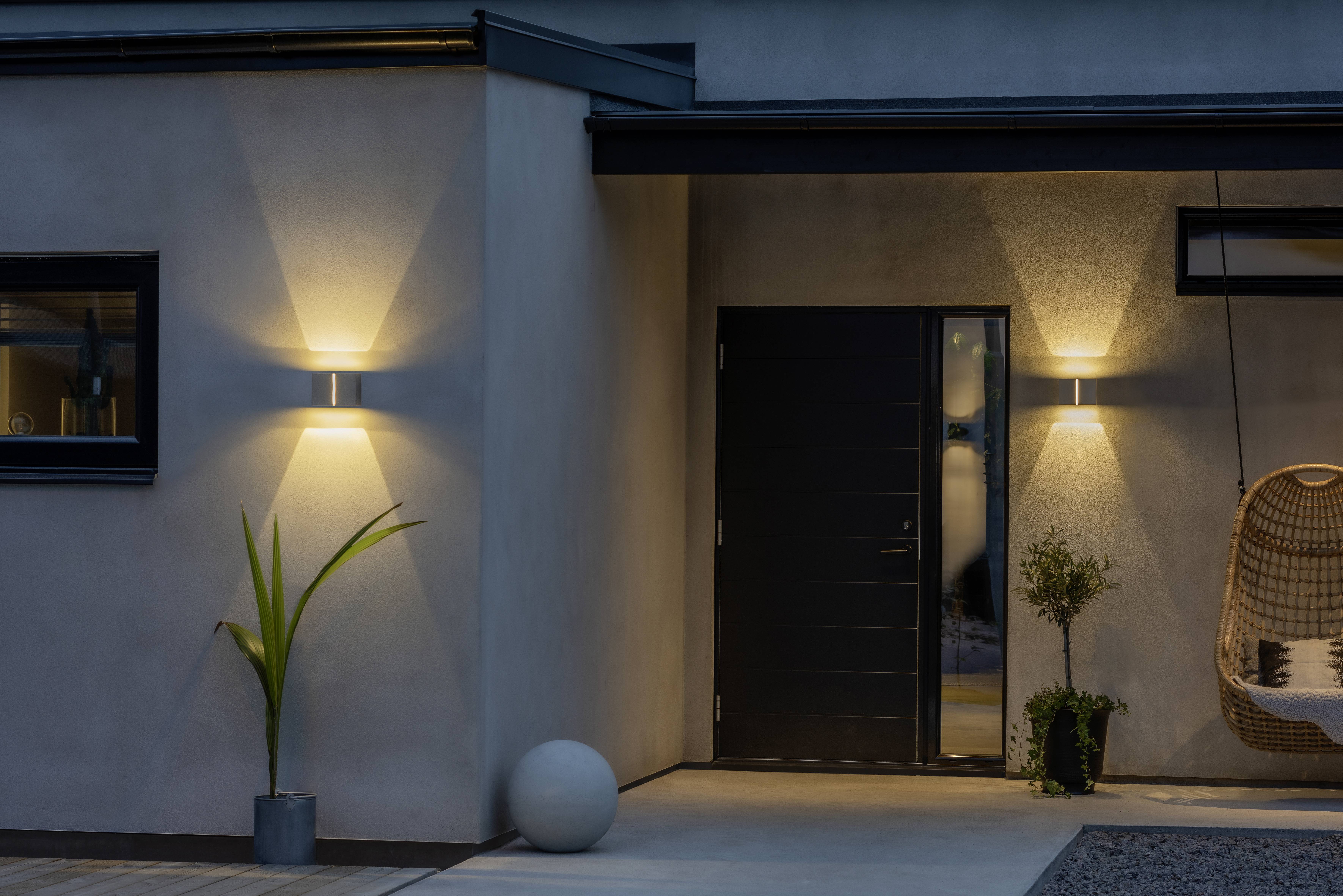 Moderna kuća s fasadom noću, osvjetljena dvjema zidnim lampama koje bacaju toplu svjetlost na zidove. Vidljiva je ljuljačka na užadima i biljke.