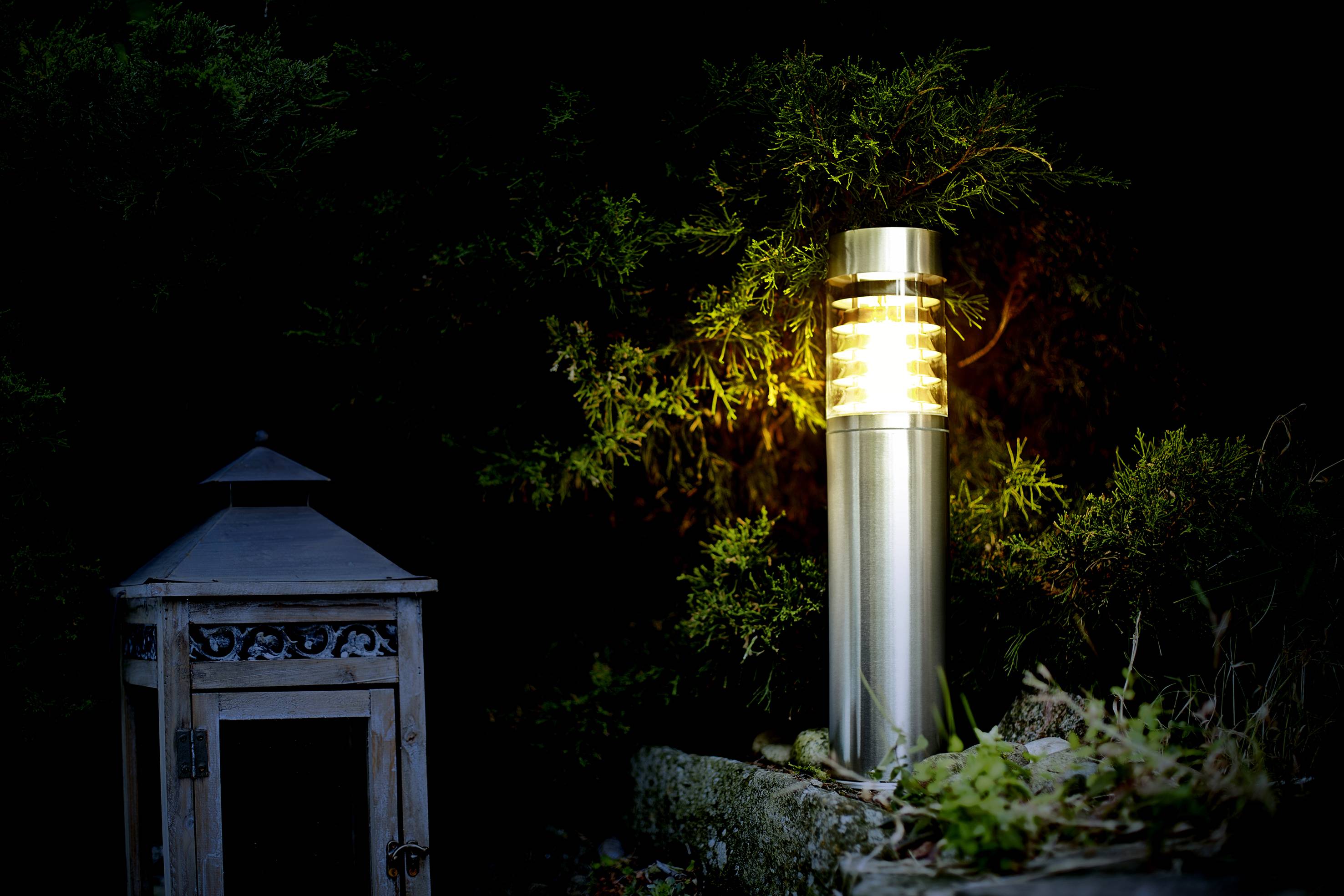 Vrtna lanterna od metala svijetli noću pokraj starog drvenog kućišta lampe, okružena tamnim lišćem.