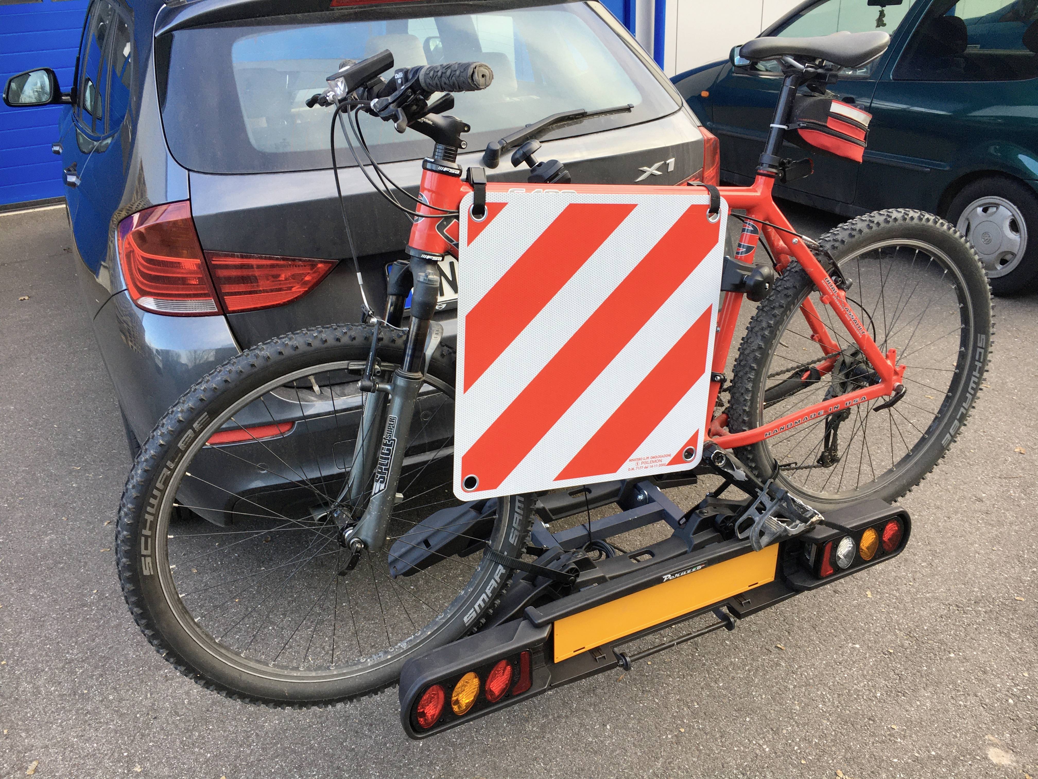 Nosač bicikla na automobilu s dva bicikla, jedan je crvene boje. Crveno-bijeli sigurnosni znak je pričvršćen na nosač.