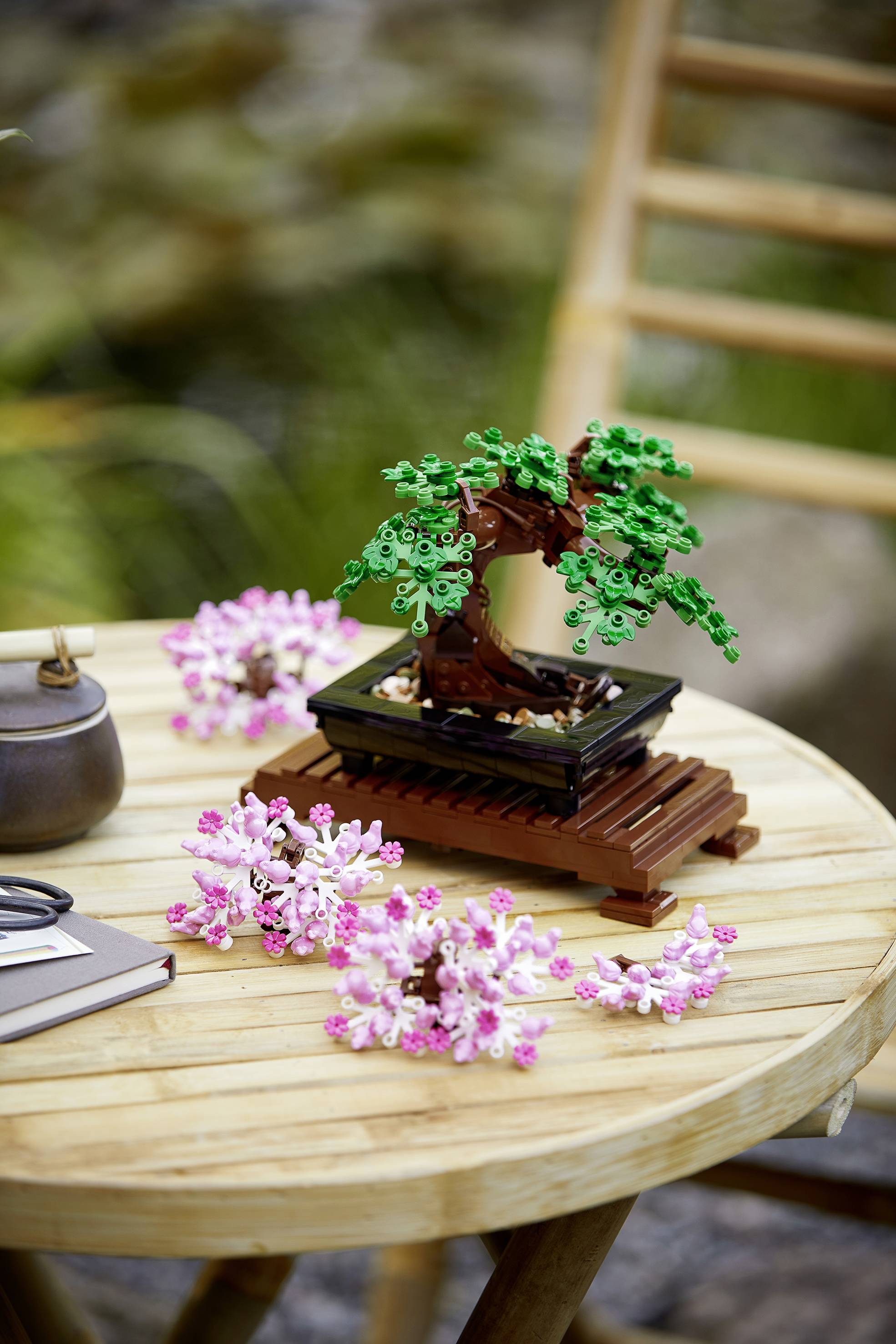 Umjetnički bonsai izrađen od kockica smješten na drvenom stolu, okružen ružičastim cvjetovima. U pozadini nejasna stolica i priroda.