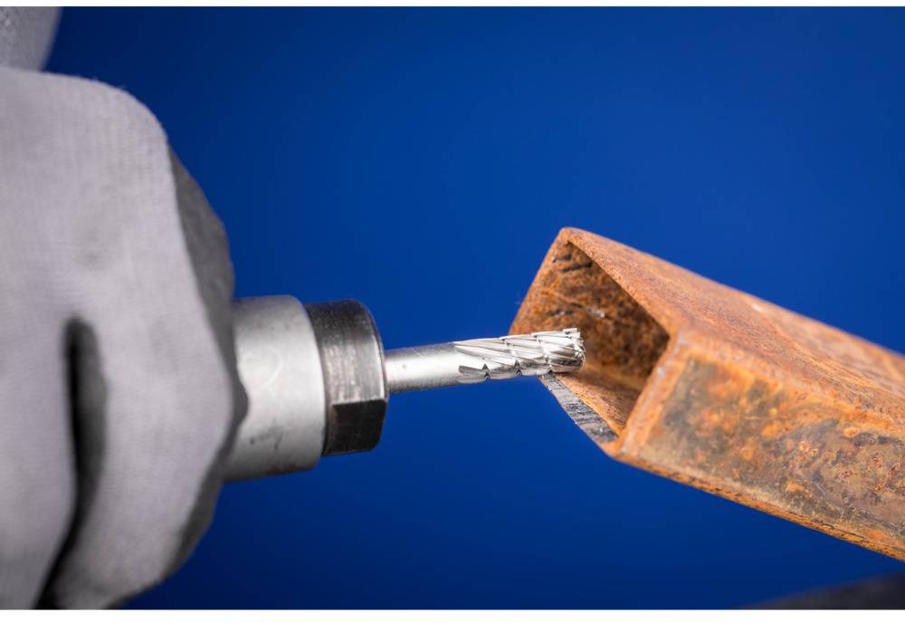 Osoba koja bušilicom obrađuje pravokutnu, hrđavu metalnu šipku. Pozadina je plava.