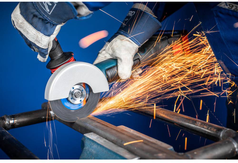 Osoba reže metal kutnom brusilicom. Varnice lete dok nosi zaštitnu opremu. Pozadina je plava.