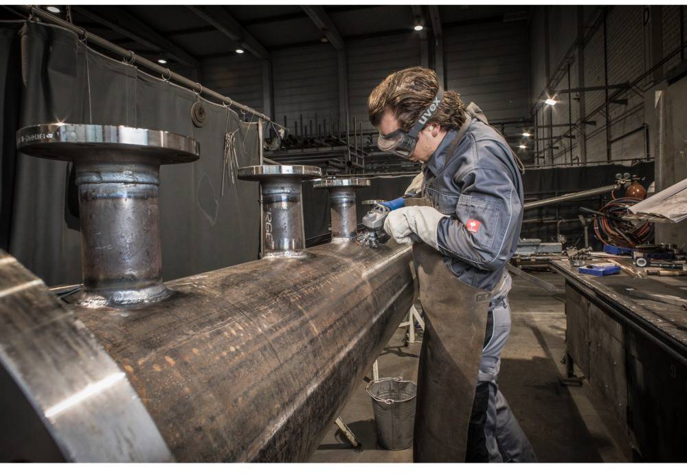 Muškarac u radnoj odjeći zavaruje metal u velikoj radionici. Nosi zaštitne naočale i rukavice, industrijski ambijent.