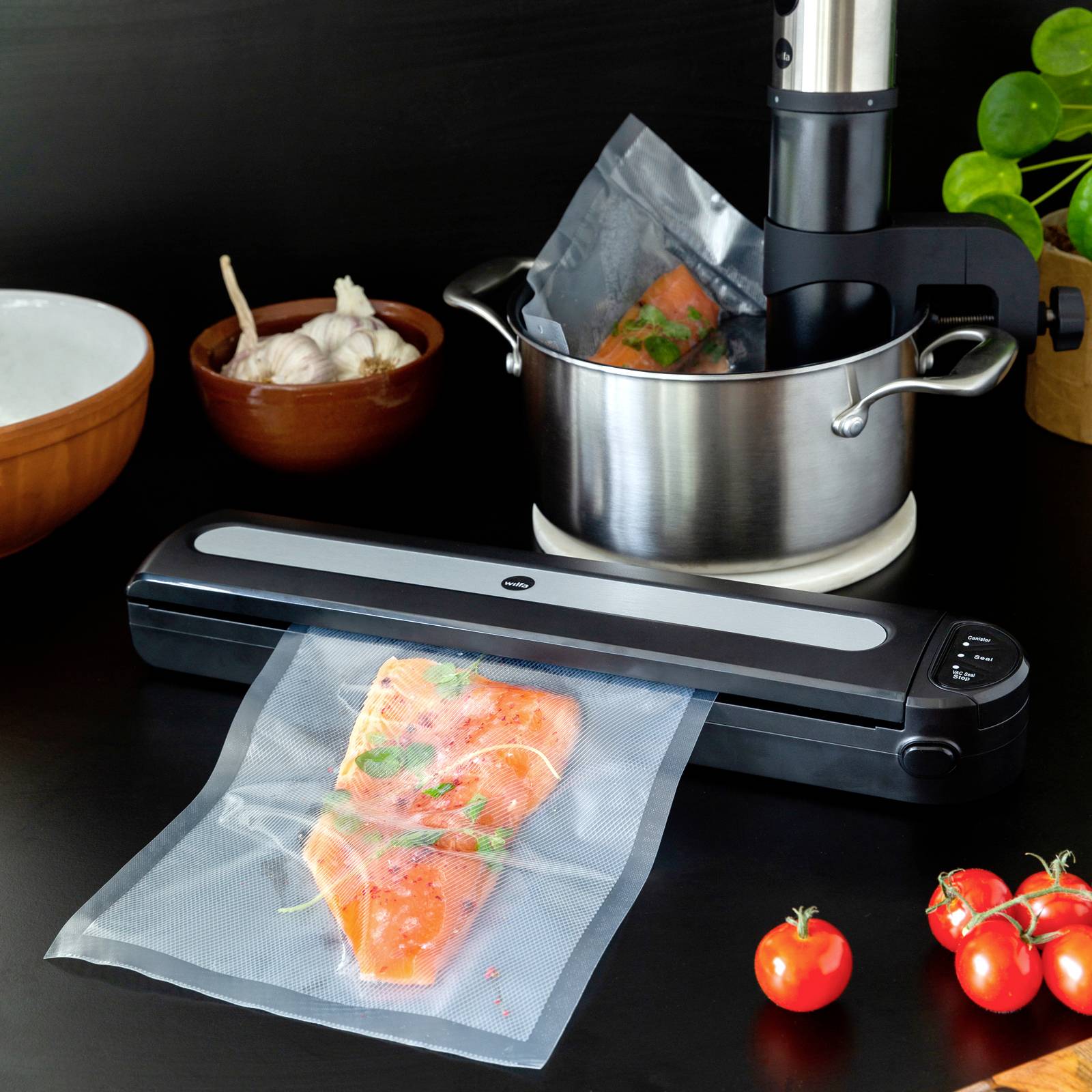 Uređaj za vakumiranje brtvari komade lososa s začinskim biljem u vrećici. U pozadini se nalaze lonac i uređaj za sous vide kuhanje te češnjak.