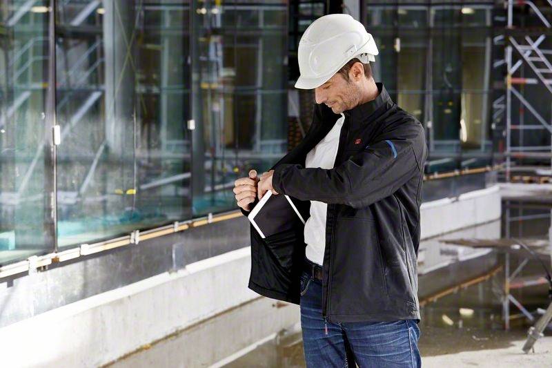 Muškarac sa zaštitnom kacigom stoji na gradilištu i gleda u otvoreni džep jakne dok drži tablet.